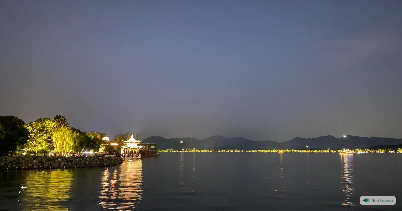 At night, a lit pavilion and willows on a point of land, lights along the far shore of a dark lake