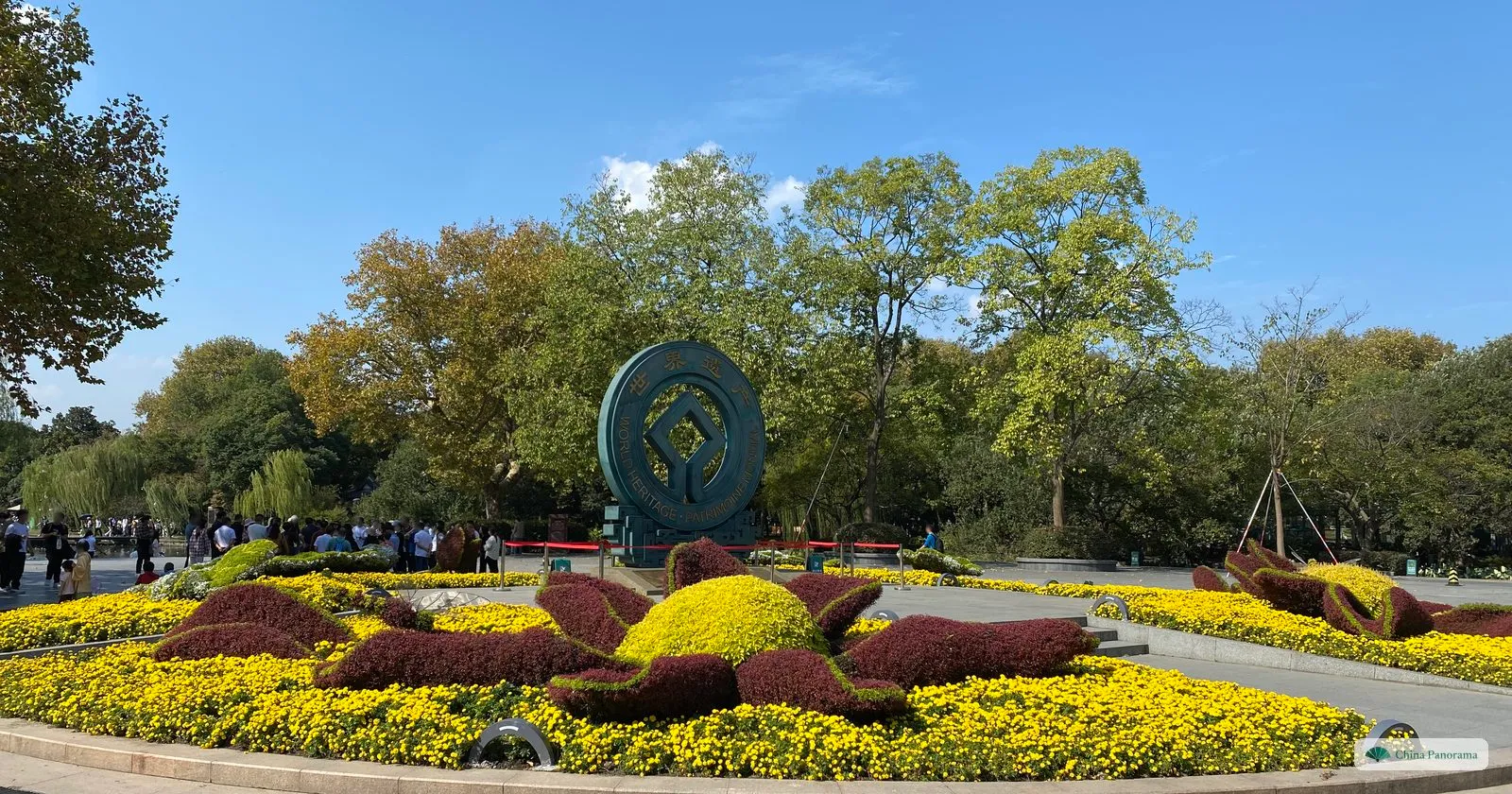 A large round bronze World Heritage emblem in flower beds of yellow chrysanthemums and red foliage, trees behind