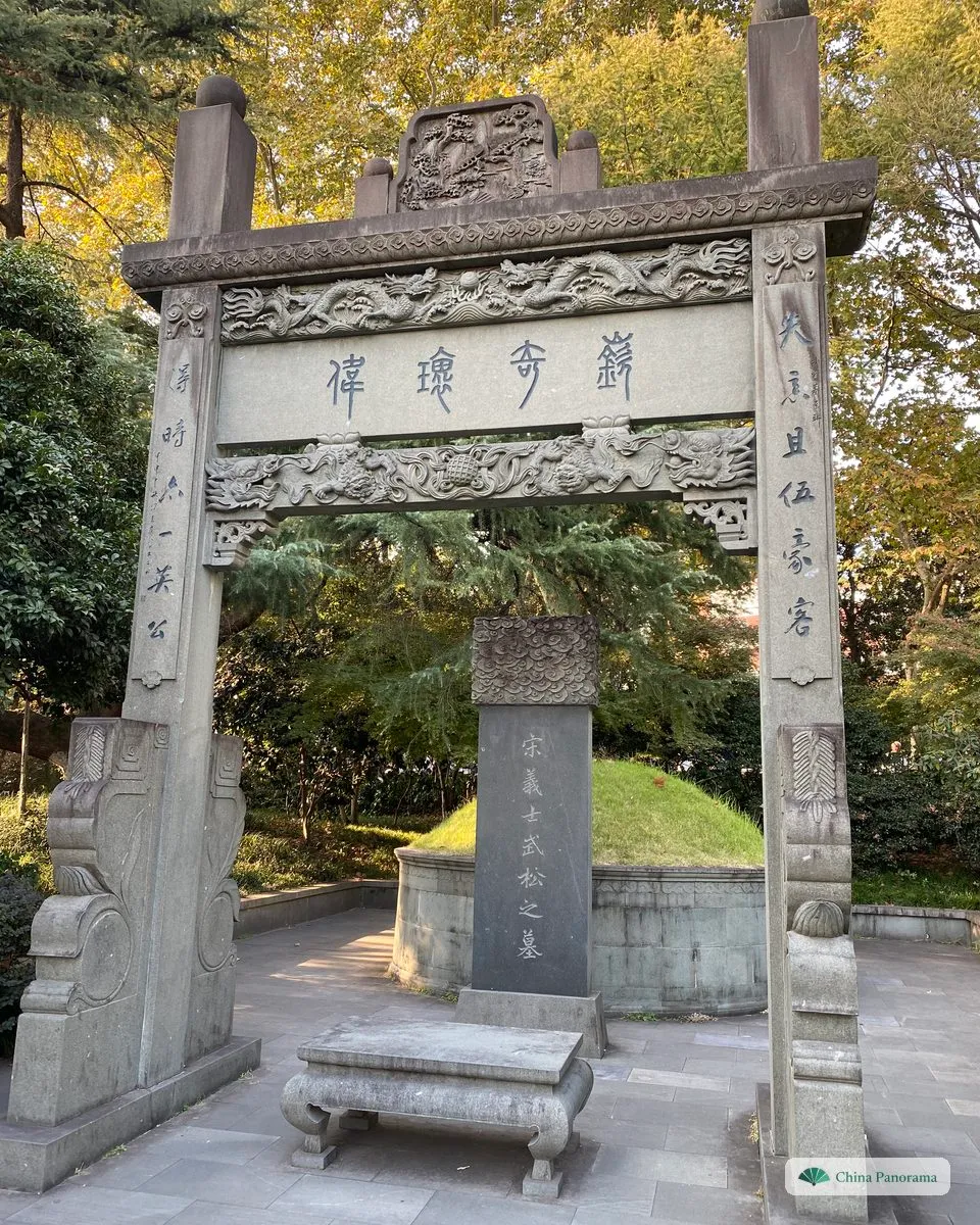 A carved stone memorial archway framing a stele and a grassy grave mound, trees behind