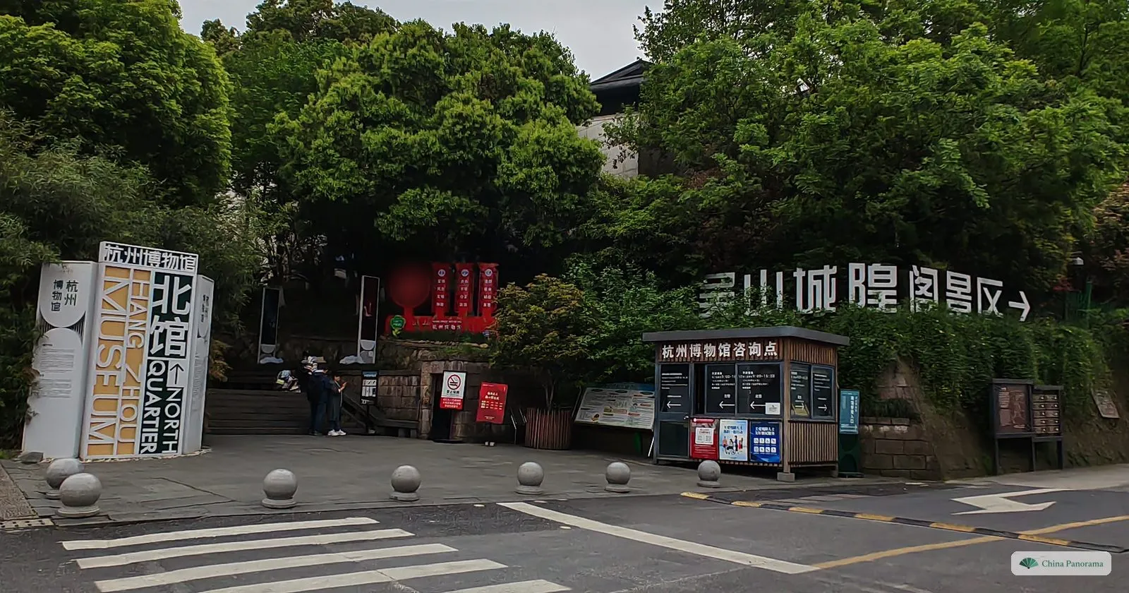 The entrance to a hillside park with large white sign boards and a small information kiosk, trees above
