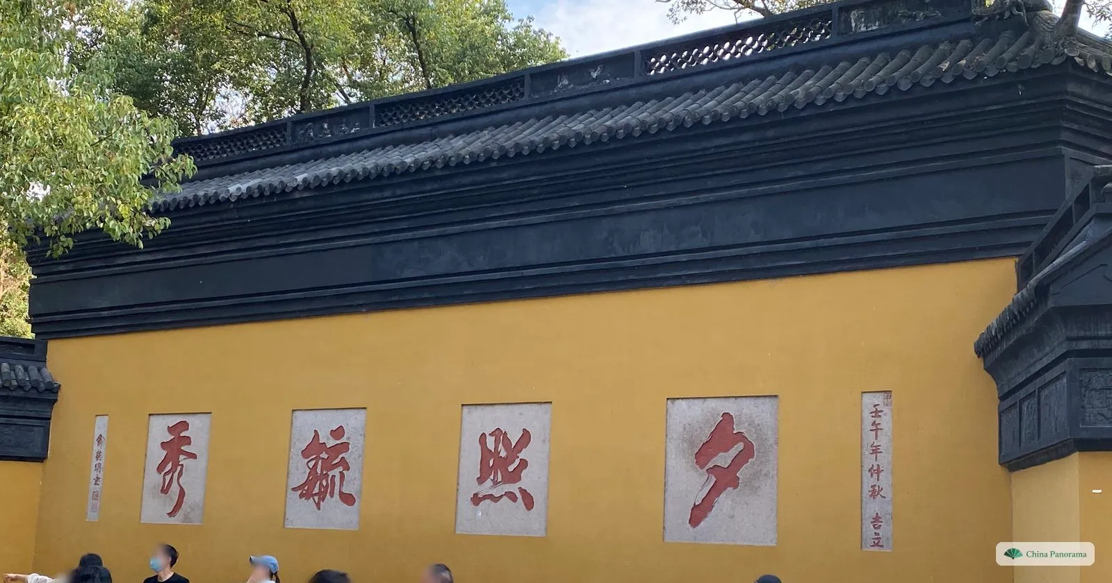 A long yellow wall under a black-tiled coping with four large red characters, visitors walking past