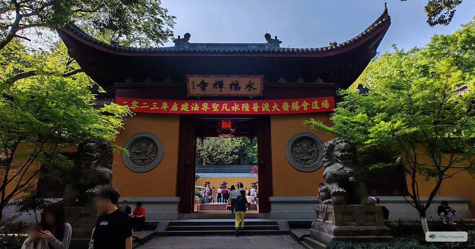 A temple gate in a yellow wall with a plaque of four characters, a red banner, two stone lions and maples