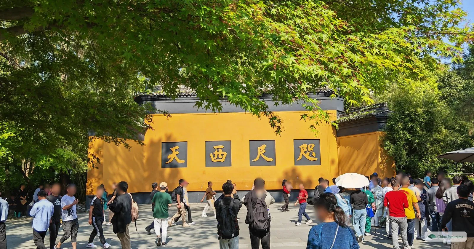 A long yellow wall with four large dark characters on a paved square, crowds of visitors walking past under maple trees