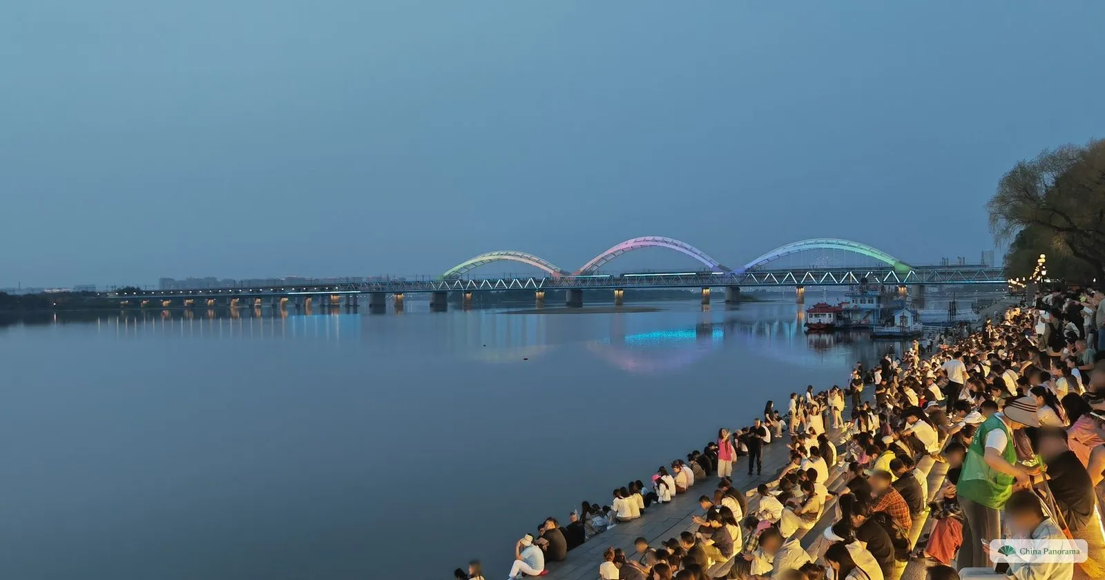 The Songhua River railway bridges lit at dusk in Harbin, with crowds sitting on the embankment steps