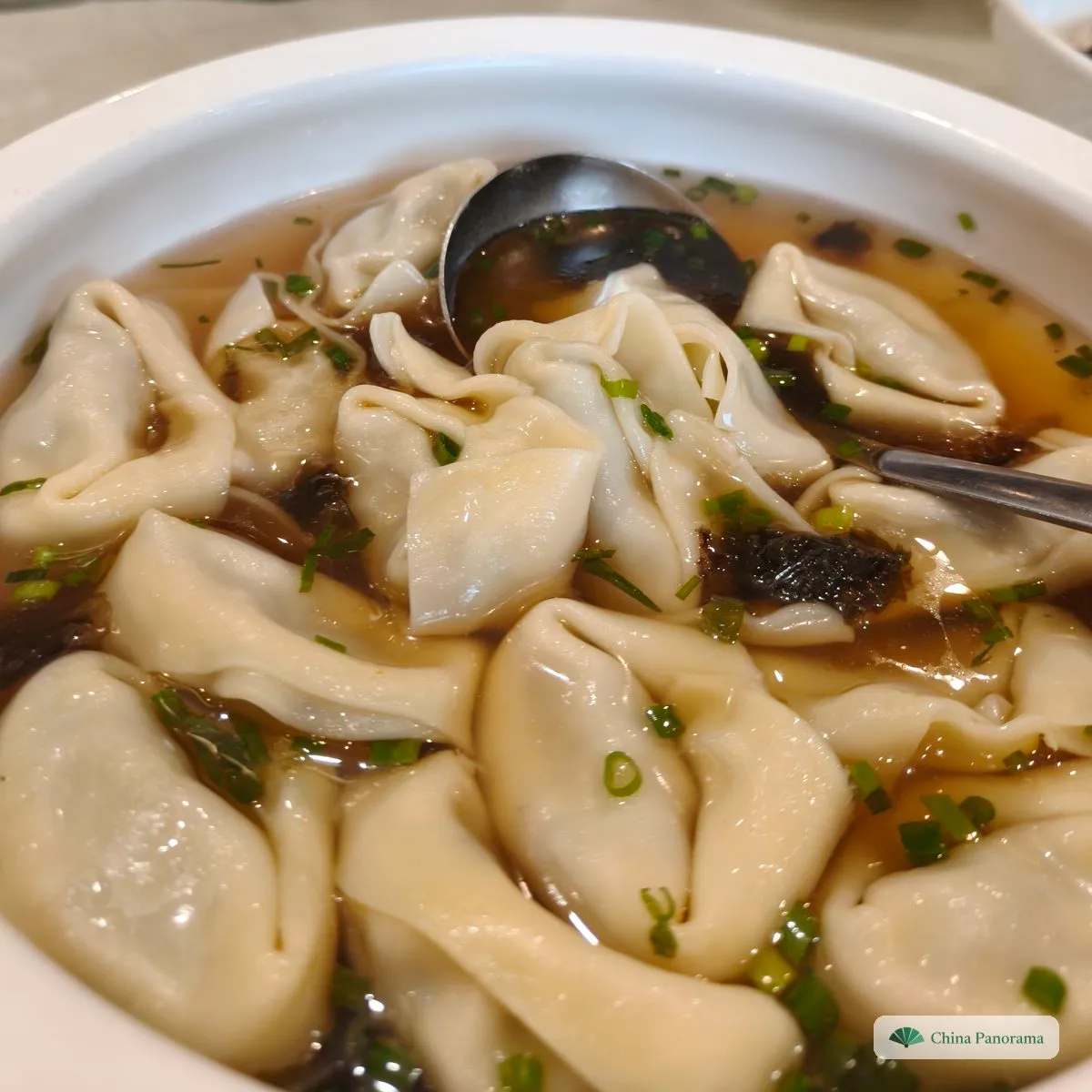 A bowl of large, loosely folded parcels with soft trailing edges in a light broth with seaweed and chopped scallions, a spoon in it