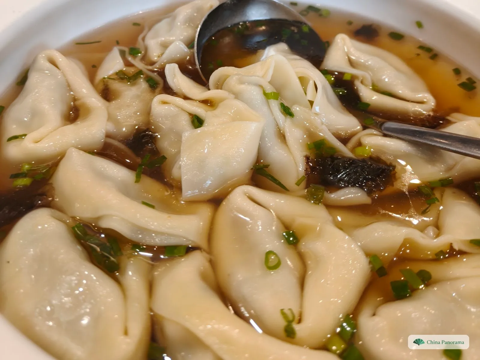 Large wontons with frilled, folded edges crowding a bowl of light brown broth with seaweed, chopped spring onion and a ladle