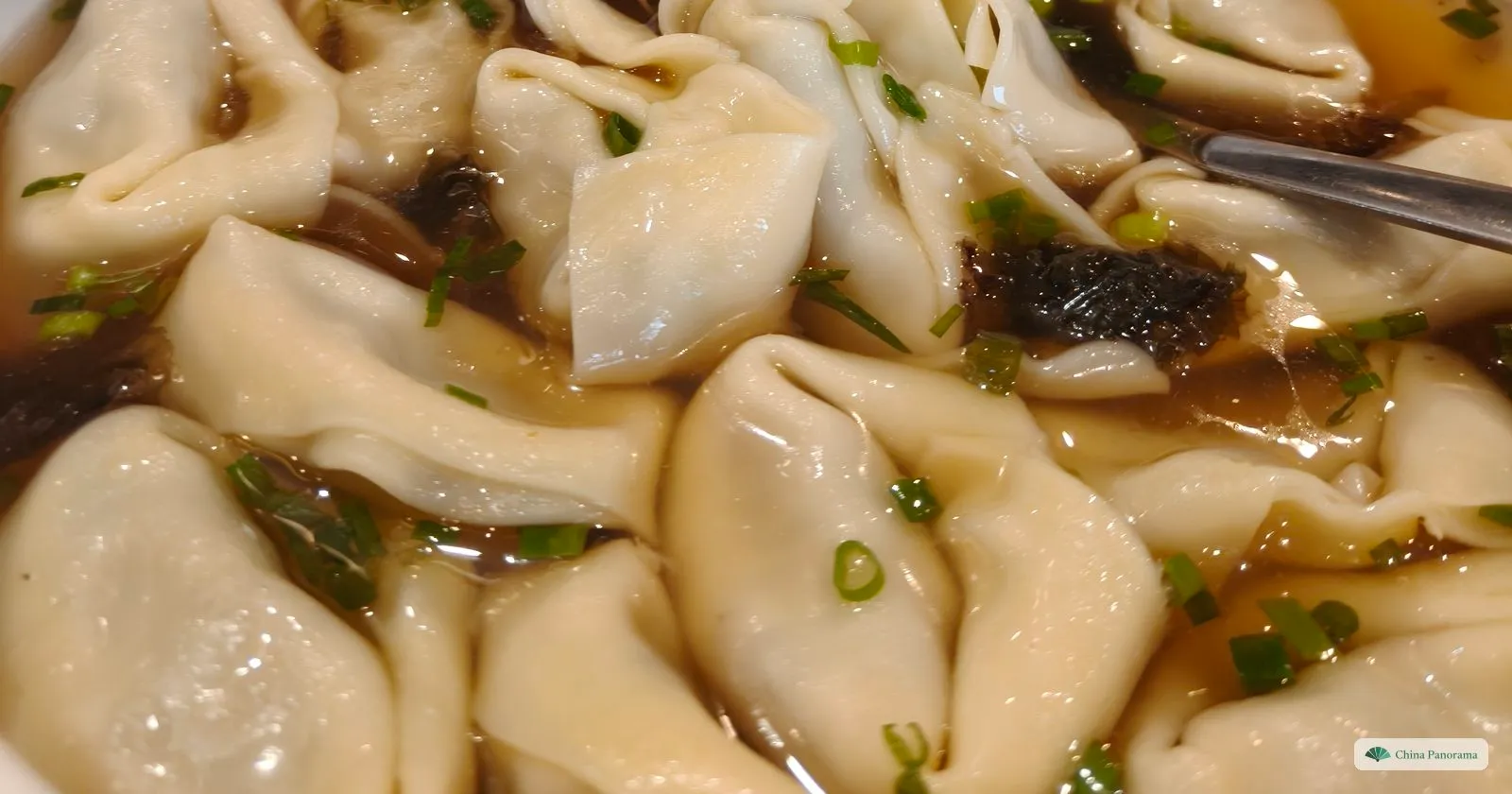 A white bowl crowded with large, loosely folded wontons with soft frilled edges in light brown broth, scattered with seaweed and chopped spring onion