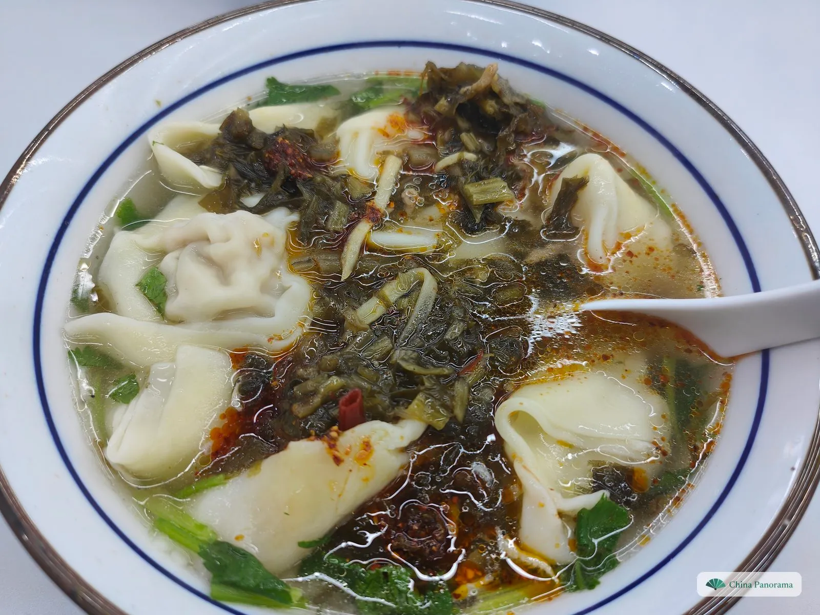 A white bowl with a blue rim: plump folded wontons in clear broth under a heap of dark pickled greens, red chilli oil and coriander leaves, a spoon at the right