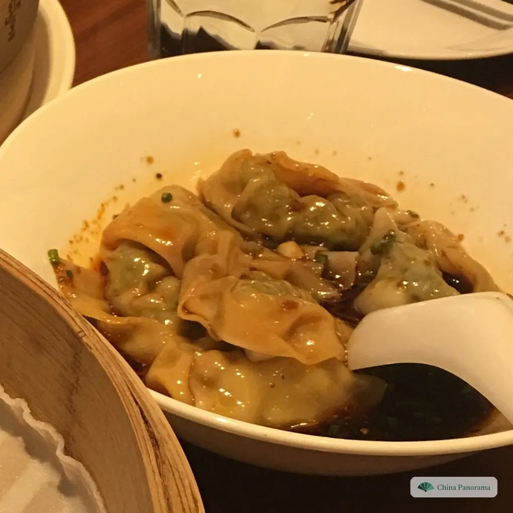 Wontons with a green filling in a pool of red chilli oil and dark sauce, a white spoon in the bowl