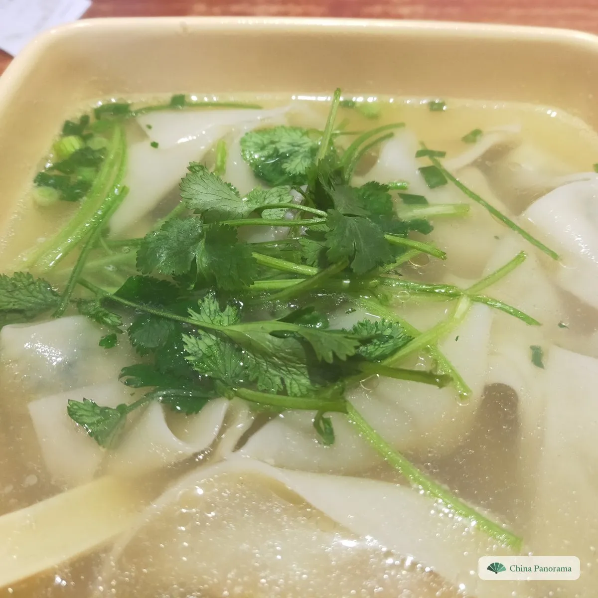 Wide, flat wontons in pale broth under a heap of fresh coriander, in a square cream bowl