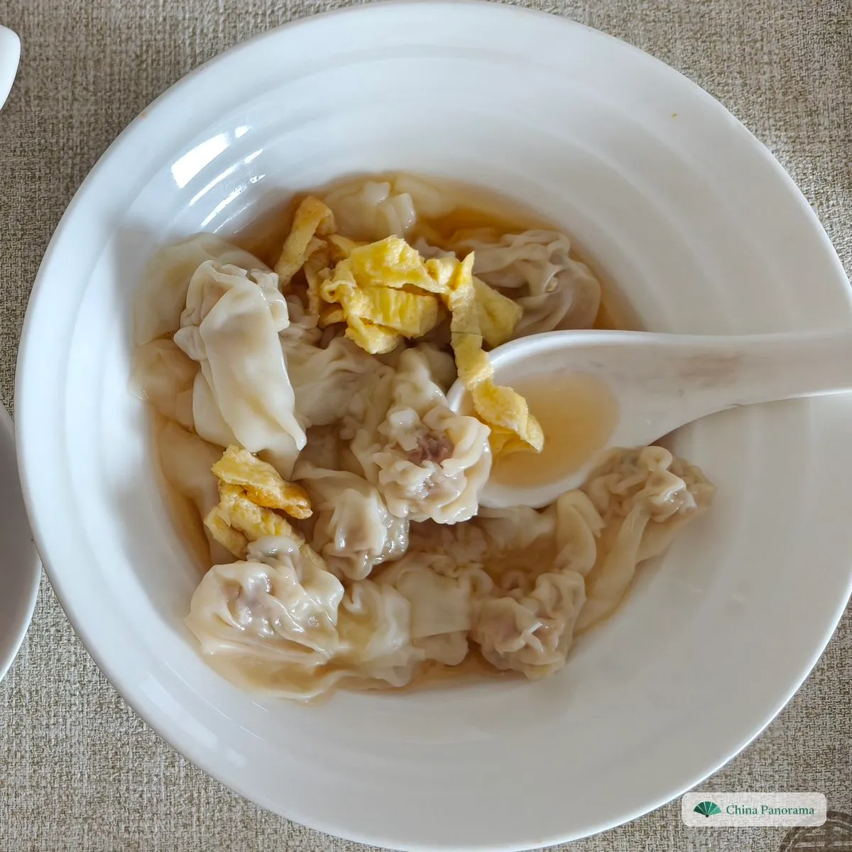 Small crumpled wontons with yellow scraps of egg crepe in a white bowl, a spoon in the broth