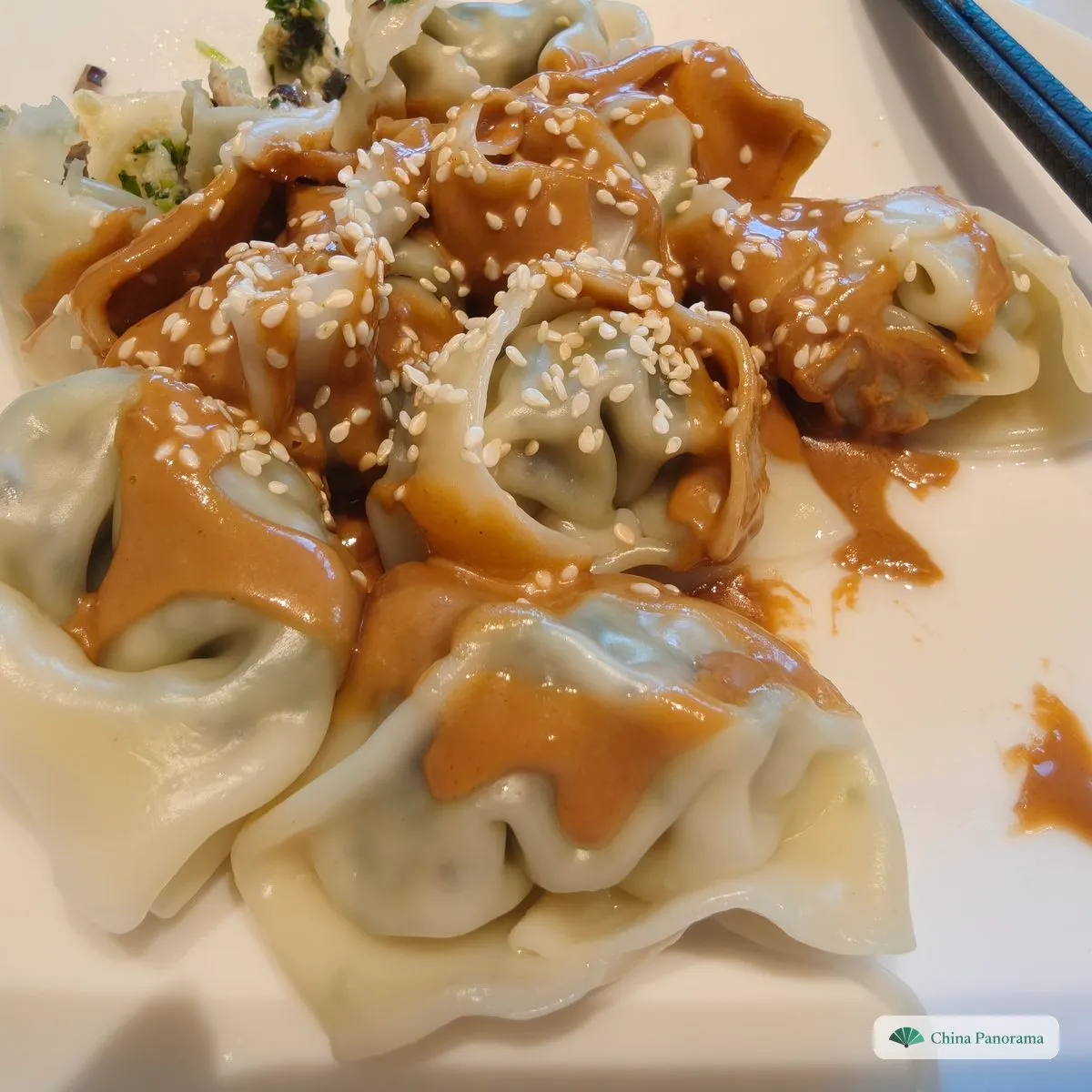 Big wontons with a green filling on a plate, without broth, covered in brown sesame sauce and white sesame seeds