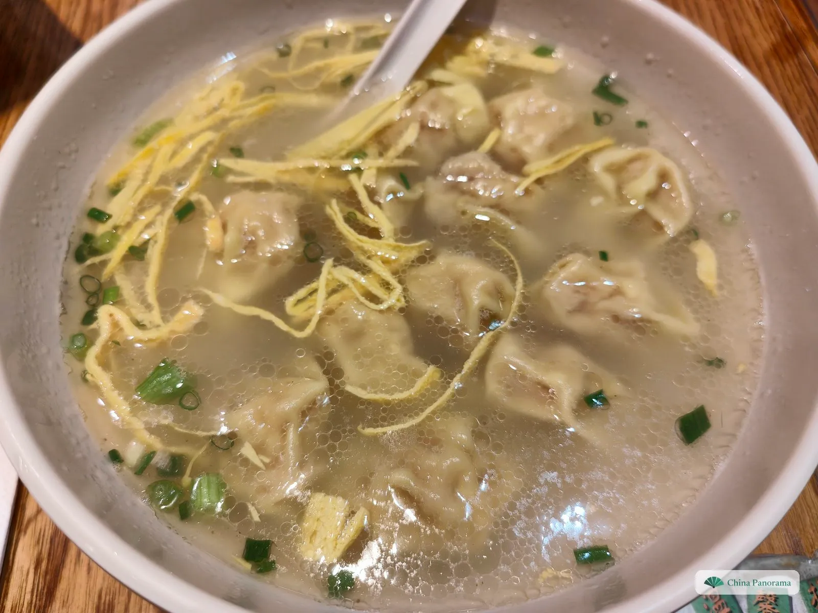 A white bowl of clear broth full of small, crumpled wontons under ribbons of yellow egg crepe and chopped spring onion, a spoon at the top