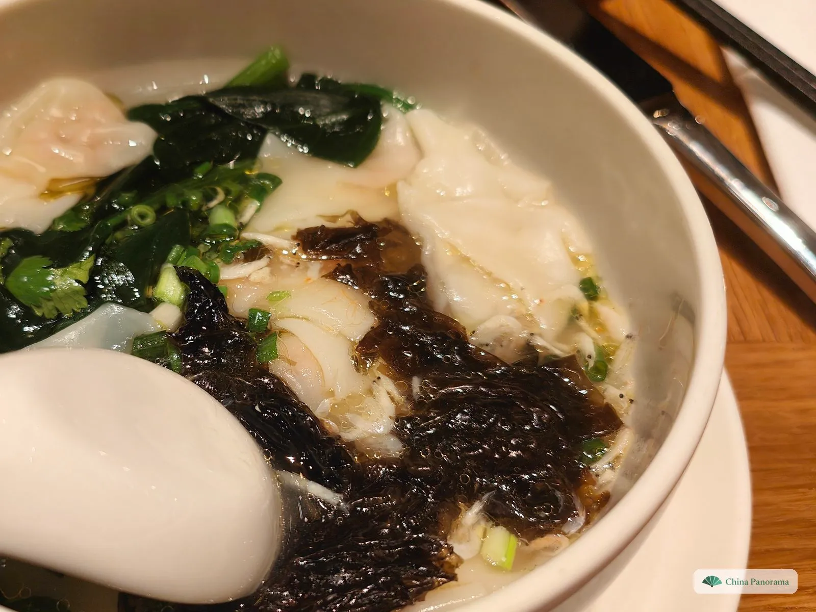 A deep white bowl with smooth, translucent wontons, dark green spinach, a tangle of black seaweed and spring onion in light broth, a porcelain spoon in front