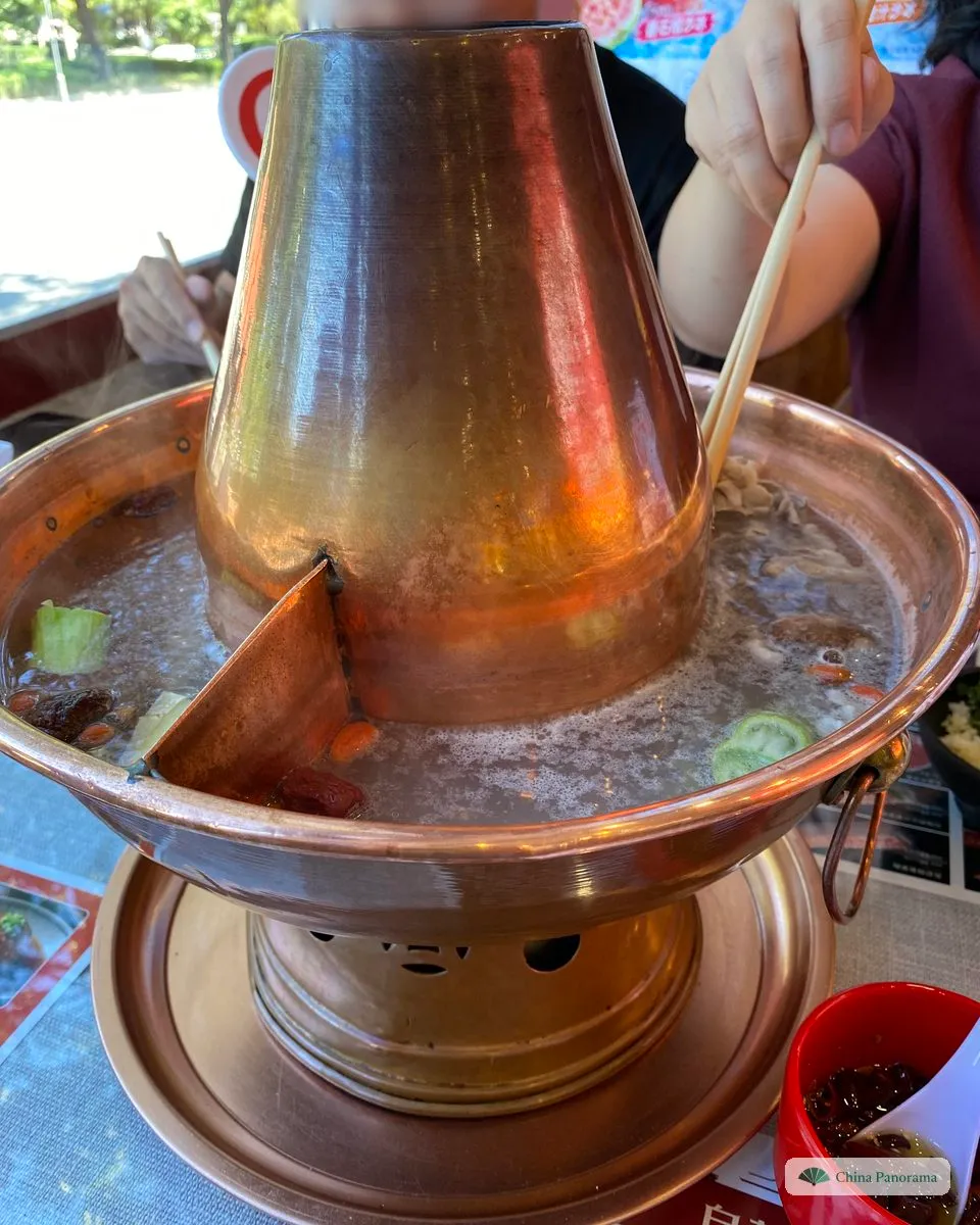 A tall copper hot pot with a chimney in the middle, clear broth with red dates, goji berries, mushrooms and vegetables around it, and a pair of chopsticks lifting cooked meat out