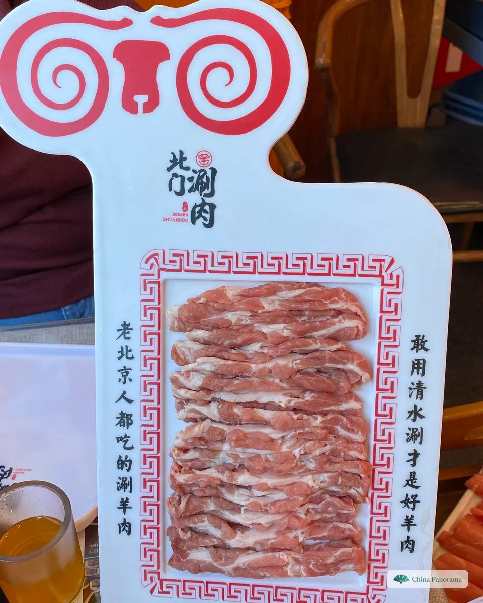 Thin raw lamb slices laid out on a white board shaped like a ram’s head, framed by a red pattern, with Chinese slogans printed down both sides