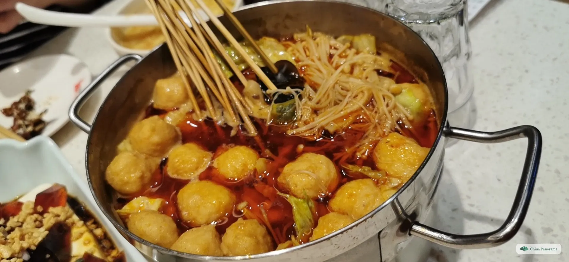 A steel pot of red chilli-oil broth full of golden balls, enoki mushrooms, cabbage and wood ear, with a bundle of bamboo skewers being lifted out; in the corner a dish of tofu with century egg
