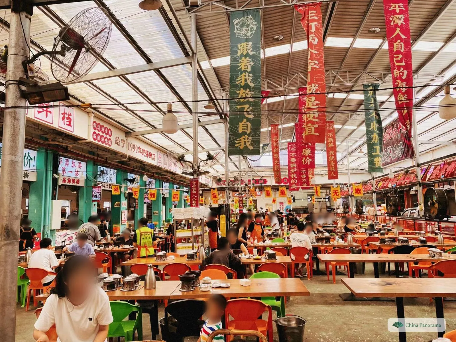 A large covered food hall with a translucent roof, long wooden tables and orange and green plastic chairs; a small pot on a burner stands at every seat, and long red and green banners with Chinese slogans hang from the roof