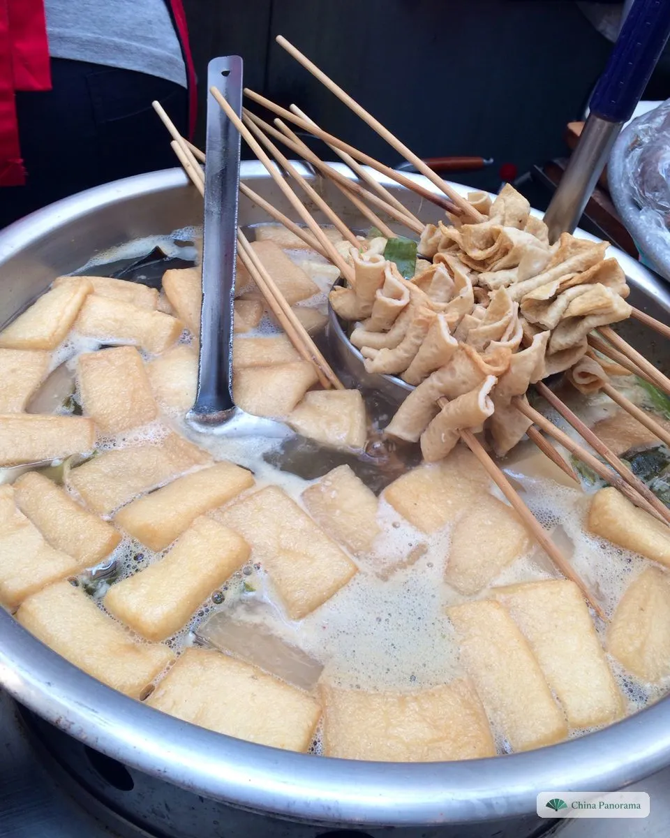 A wide steel pot at a street stall filled with golden fried fish cakes, pieces of white radish and kelp in a light broth, with a ladle and dozens of bamboo skewers of folded fish cake resting on the rim
