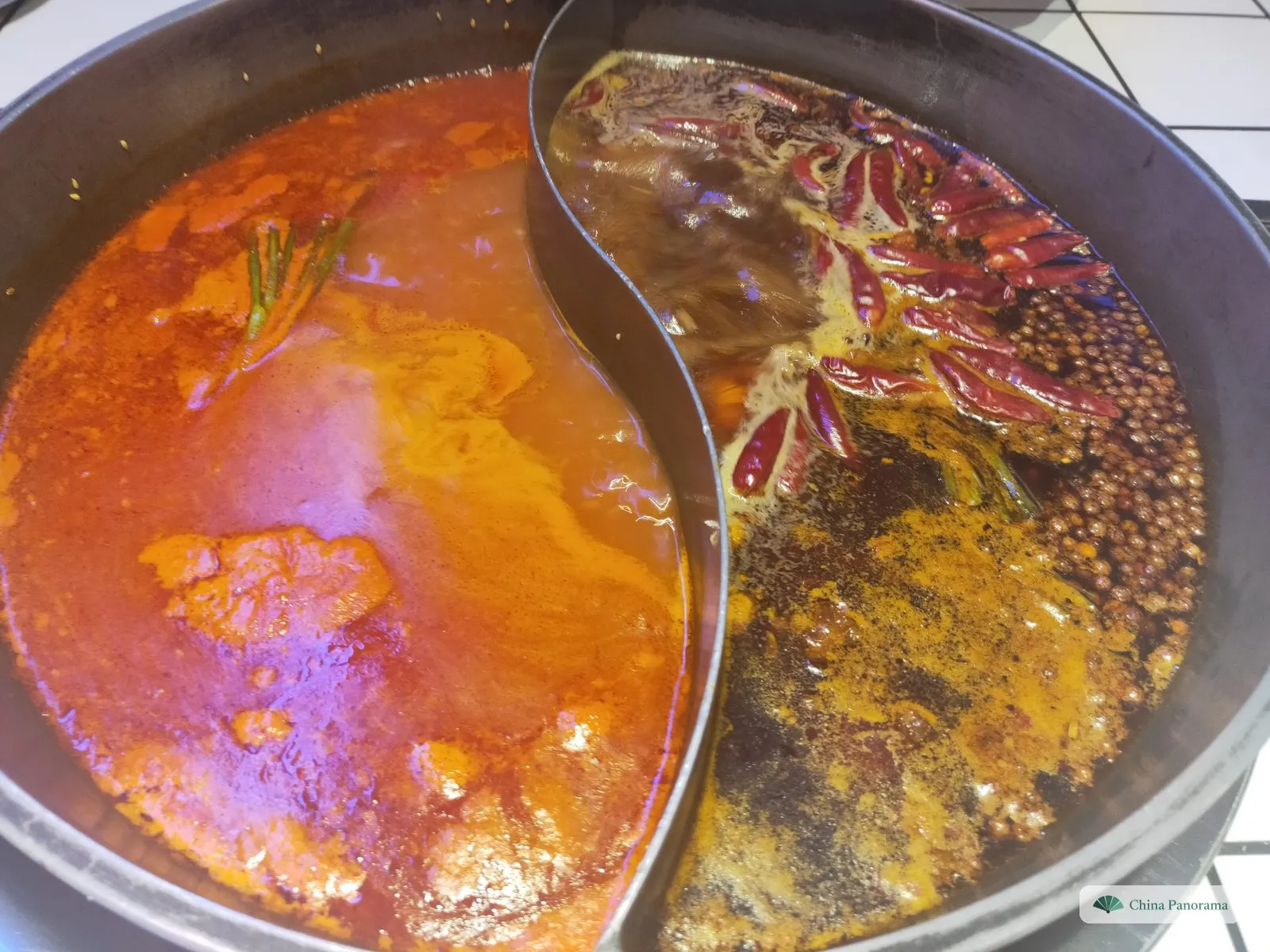 Close-up of a split hot pot: on the right, oily dark broth with whole dried red chillies and a heap of brown Sichuan peppercorns; on the left, an orange-red broth; a curved steel wall between them