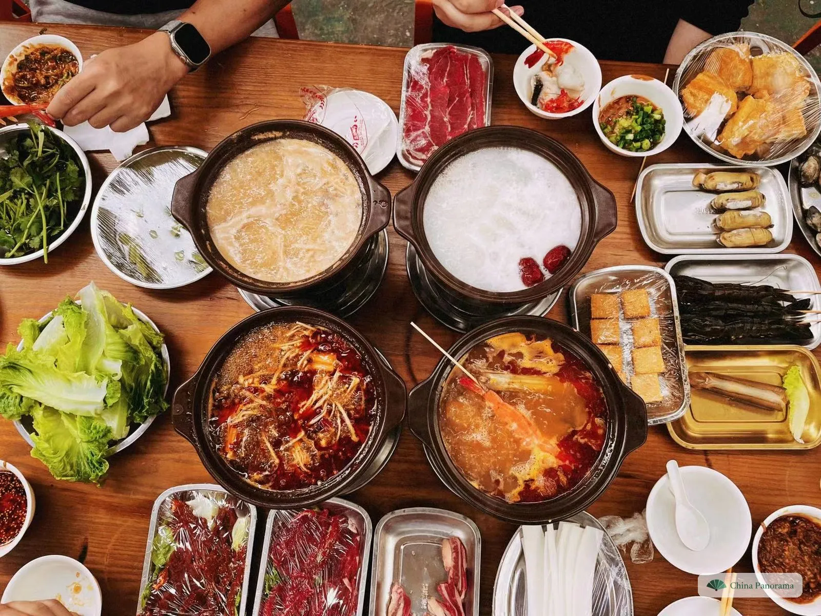 Top view of a wooden table with four small clay hot pots, each on its own burner: a pale shacha broth, white rice congee with two red dates, a red chilli-oil broth with enoki mushrooms and an orange seafood broth with a prawn on a skewer; around them plates of raw beef slices, razor clams, skewered prawns, fish tofu, lettuce and coriander