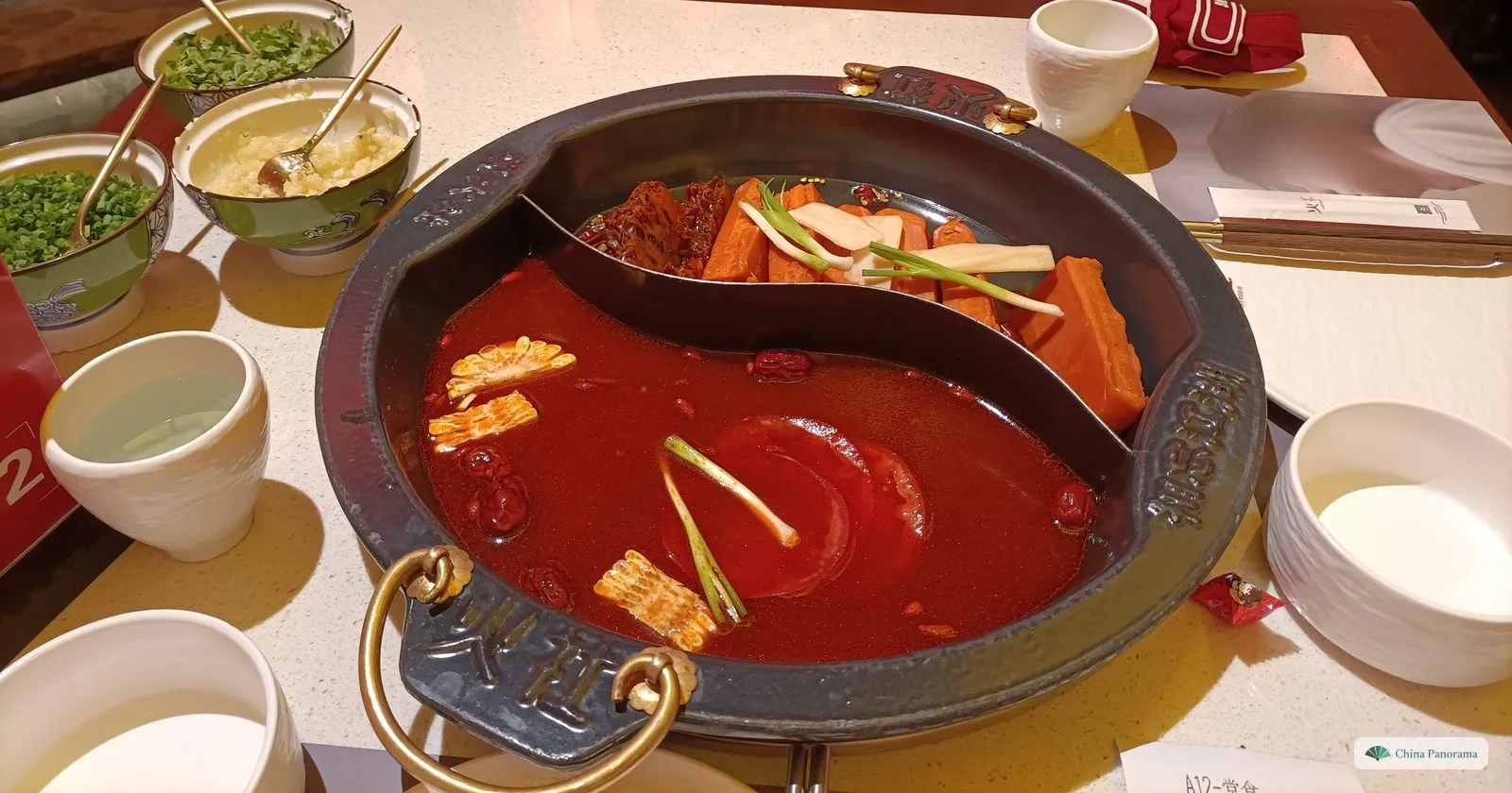 A round black hot pot split by a curved metal wall: red tomato broth with tomato slices, red dates and scallion in front, pieces of carrot and yam behind, with small bowls of chopped scallions and minced garlic and white cups on the table
