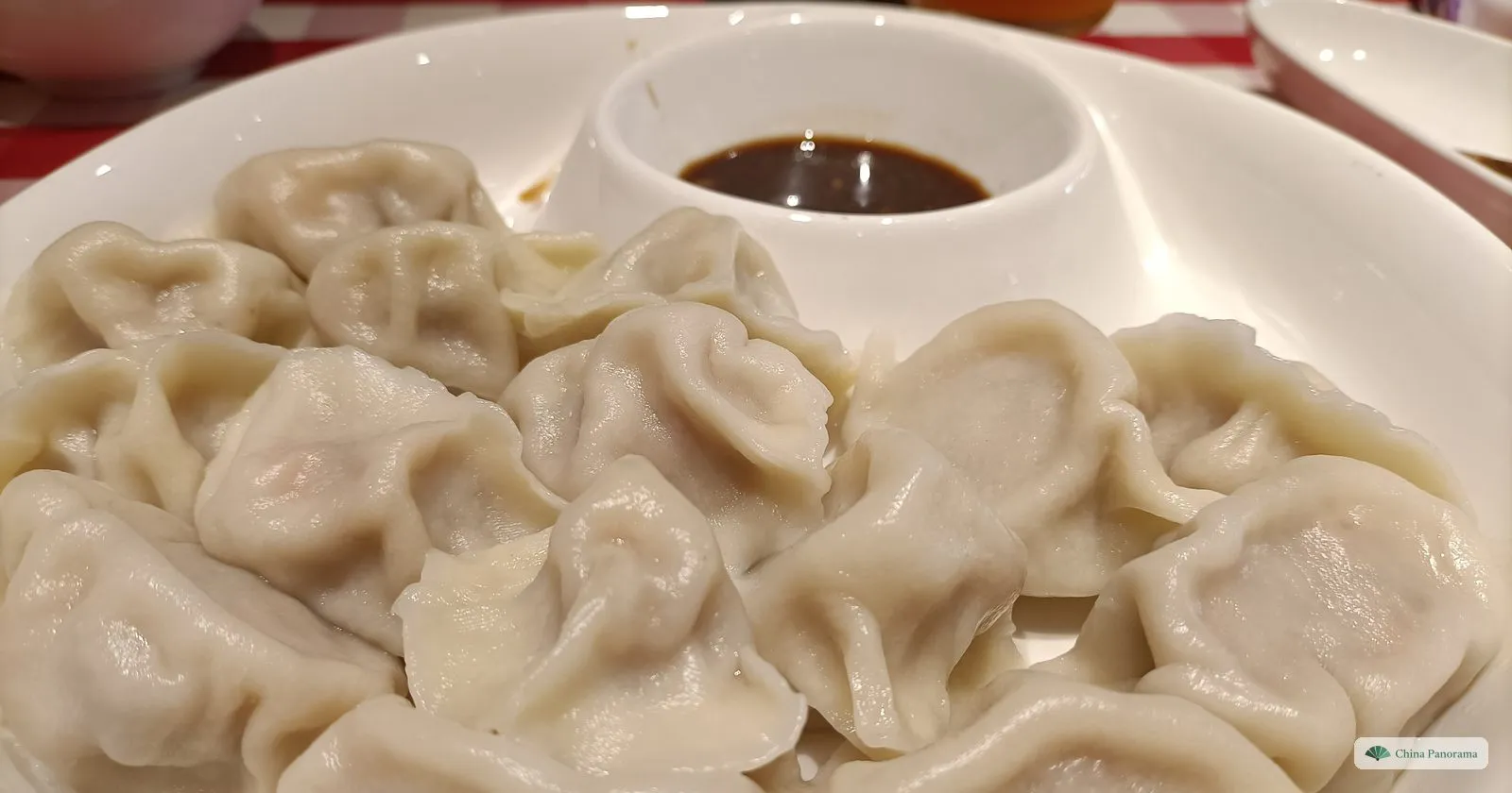 A white plate heaped with pale boiled dumplings, their pleated edges up, around a small built-in dish of dark vinegar; a red-and-white checked tablecloth behind