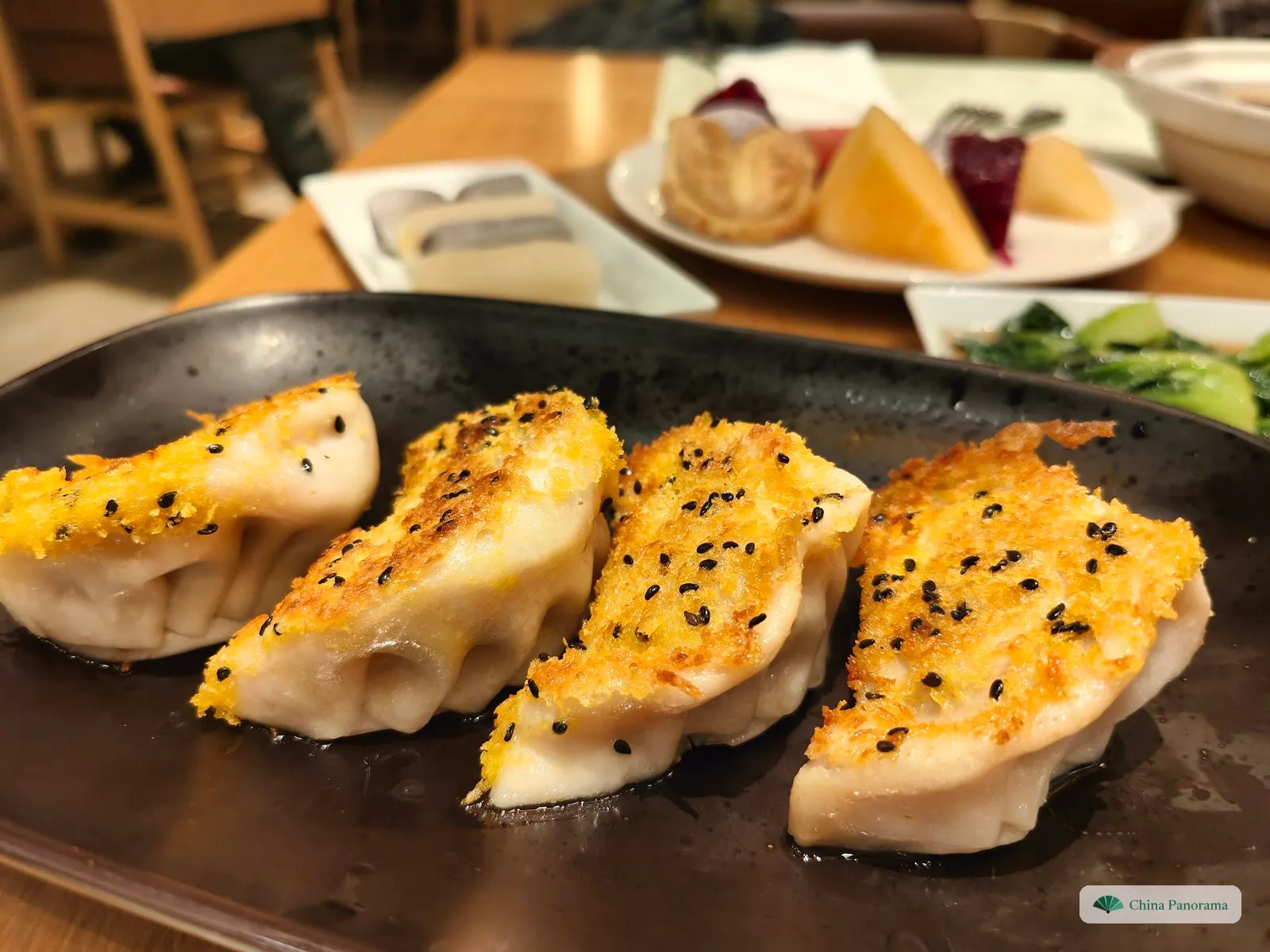 Four pan-fried dumplings in a row on a dark plate, each topped with a golden, crumbly crust and black sesame seeds; plates of fruit and greens blurred behind