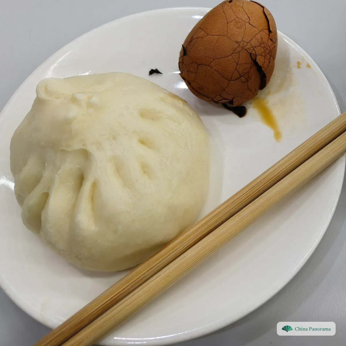 A single large, fluffy white bun with pleats twisted on top, a tea egg and chopsticks beside it on a plate