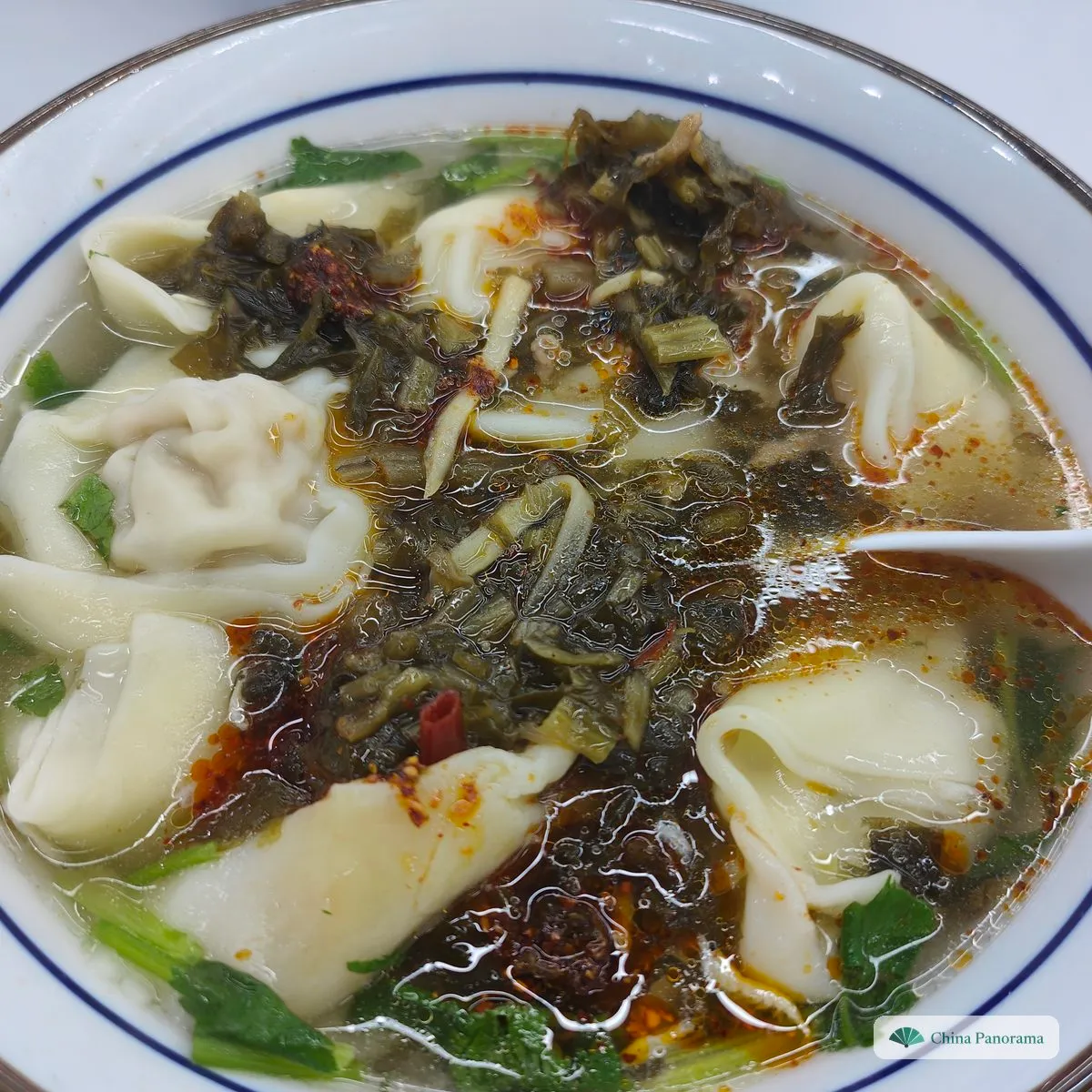 Large, loosely folded parcels in a soup with dark chopped pickled greens, red chilli oil and leafy vegetables, in a blue-rimmed bowl