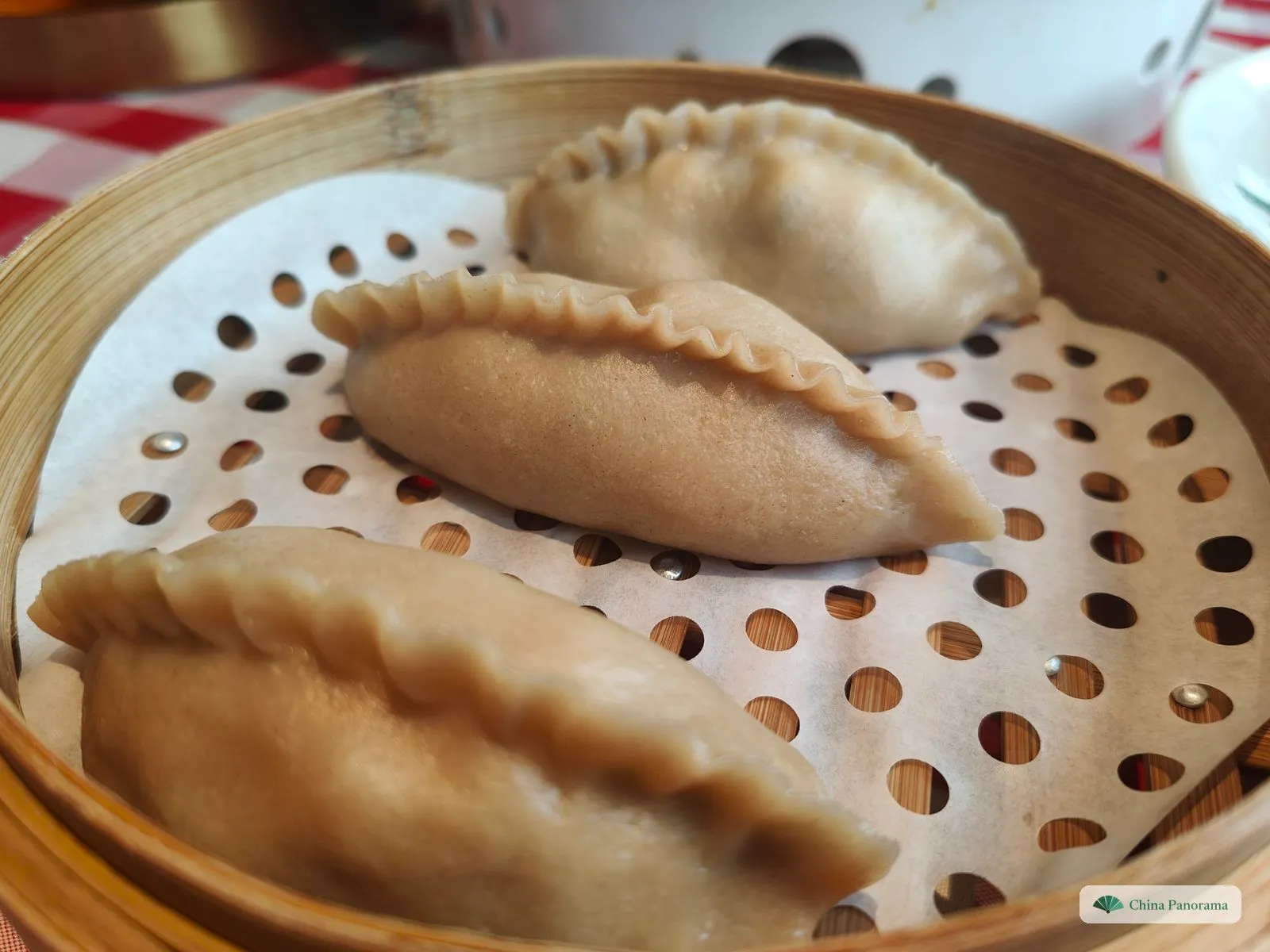 Three large light-brown dumplings with crimped, rope-like edges on perforated paper in a round bamboo steamer, a red-and-white checked tablecloth behind