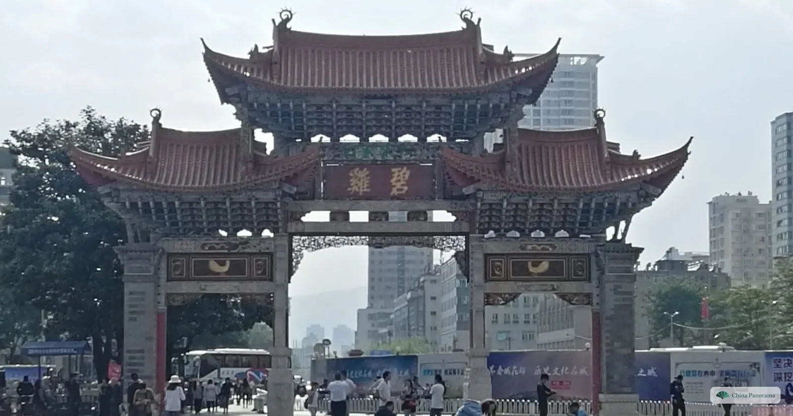 A dark wooden archway with three tiers of roofs over a busy square, buses and banners behind