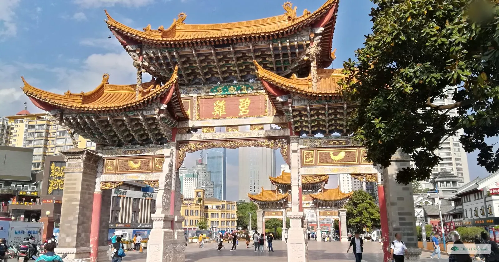 A painted archway with yellow roofs and green-and-gold panels, a second archway visible through it, a tree on the right