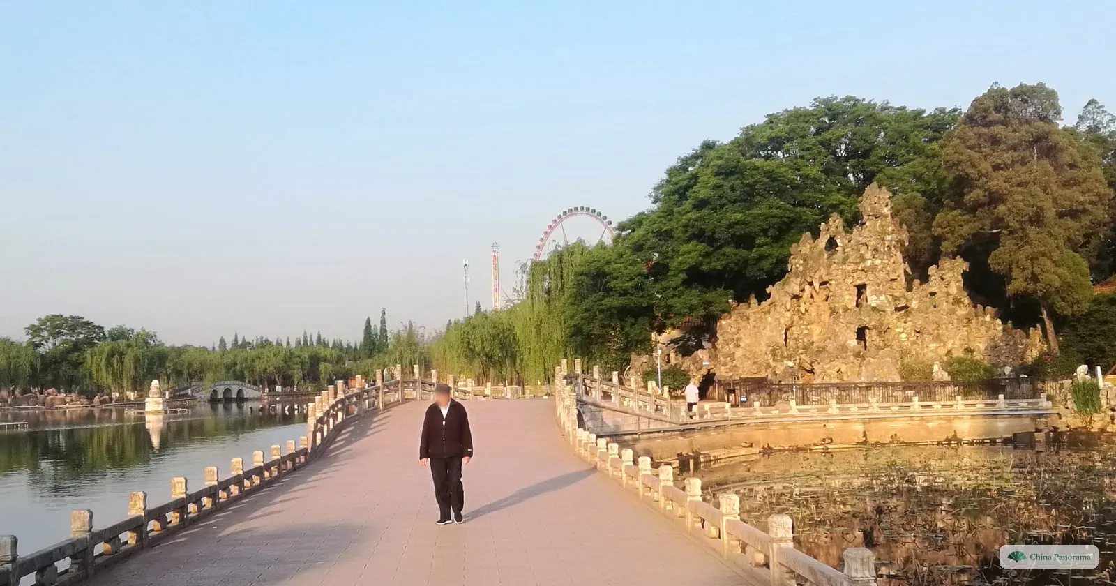 A man walking along a balustraded causeway between ponds, a rockery and a Ferris wheel in the background