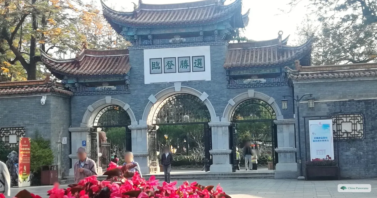 A grey-brick gateway with three arches and a white plaque of four green characters, red flowers in the foreground and people walking in