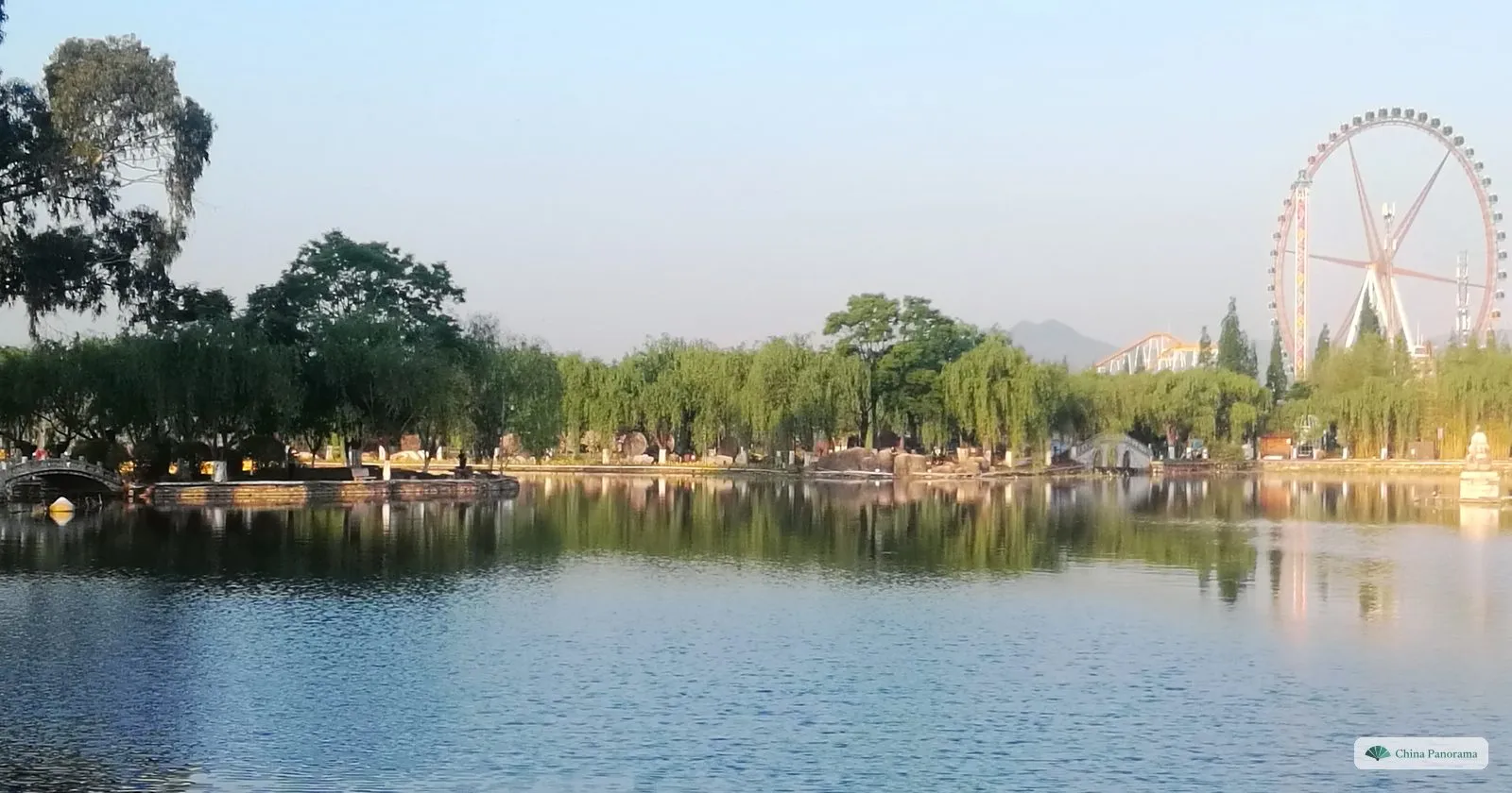 A wide calm lake edged with willows, a Ferris wheel and apartment towers on the far shore
