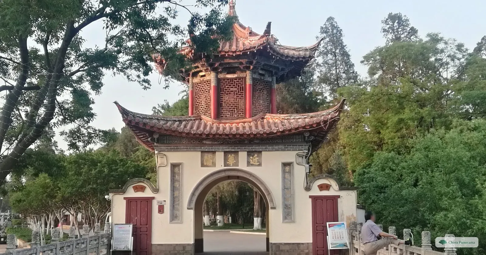 An arched gateway in a white wall with a small two-tiered pavilion on top and a plaque of three characters