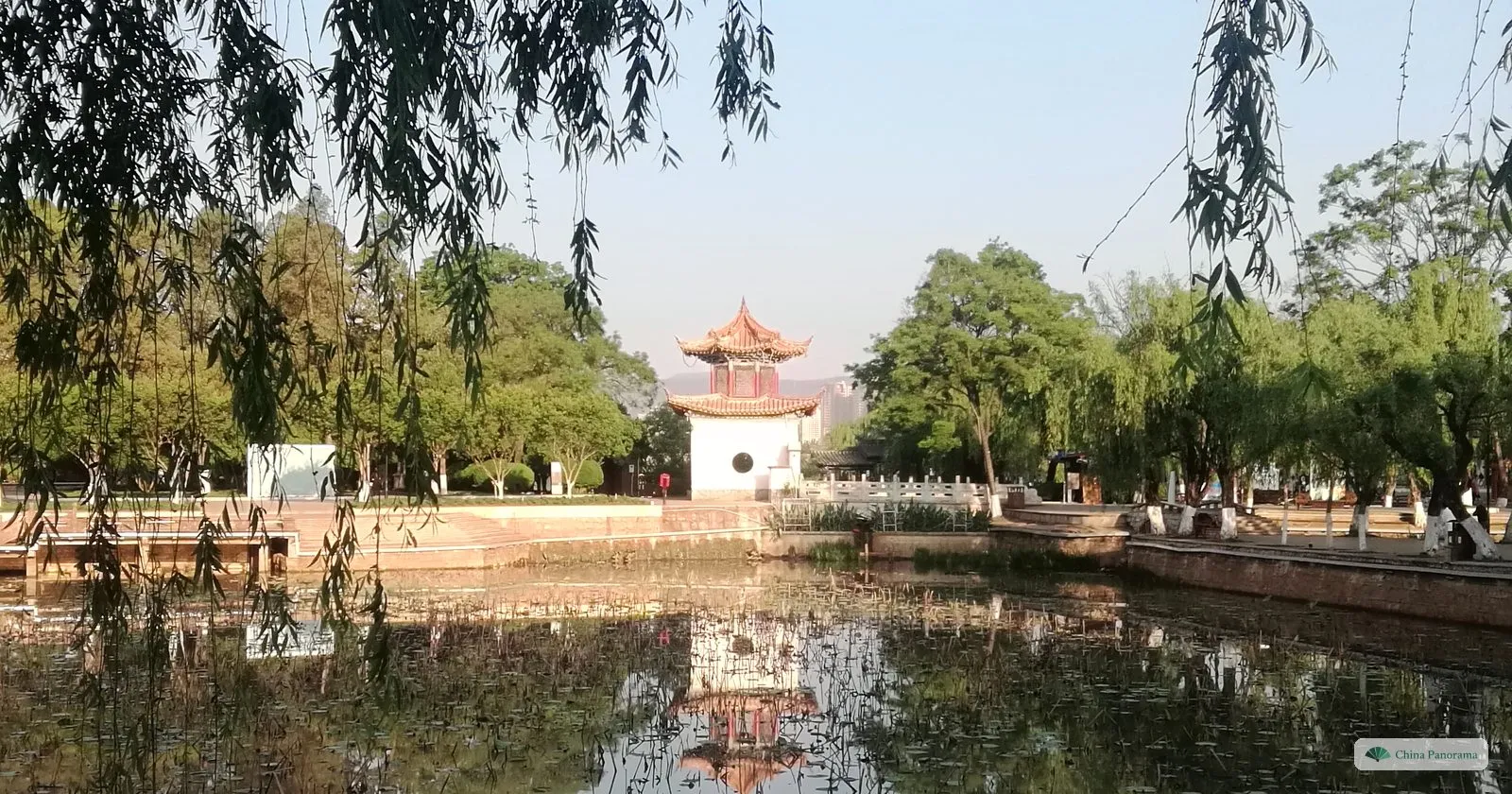 A small pavilion with a white base and orange roof across a pond, willows on either side, the scene reflected