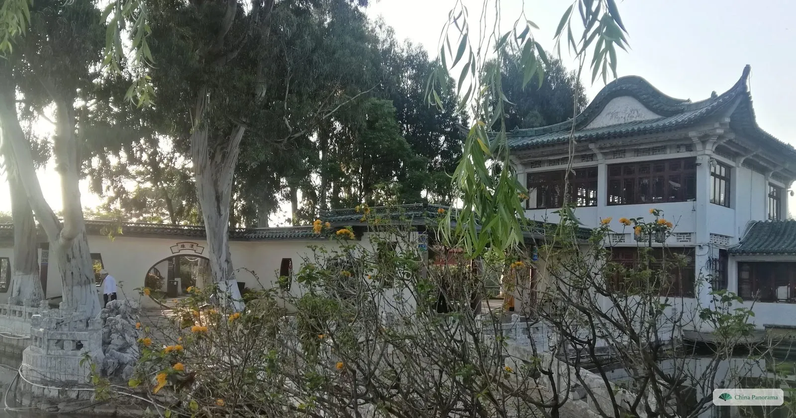 A white two-storey hall with a curved grey roof behind trees and a stone balustrade