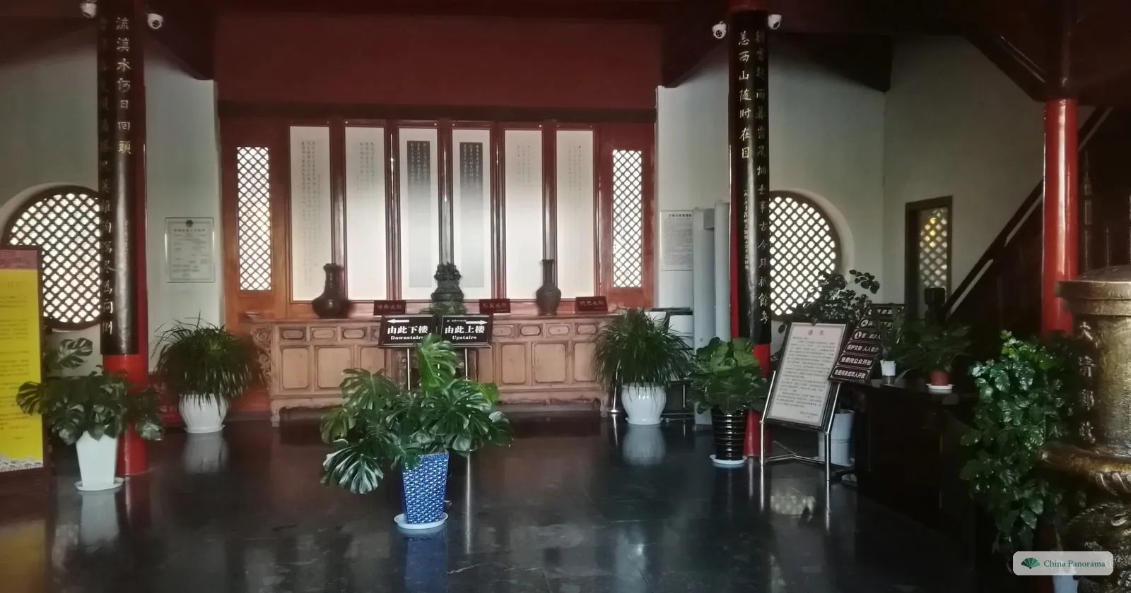 Inside a pavilion: dark pillars carved with calligraphy, a long wooden cabinet with vases under scroll panels, potted plants and signs for upstairs and downstairs