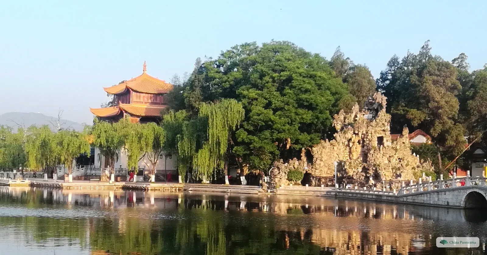 A three-storey pavilion and a large craggy rockery lit by early sun beside a still pond, an arched stone bridge on the right and a hill on the horizon