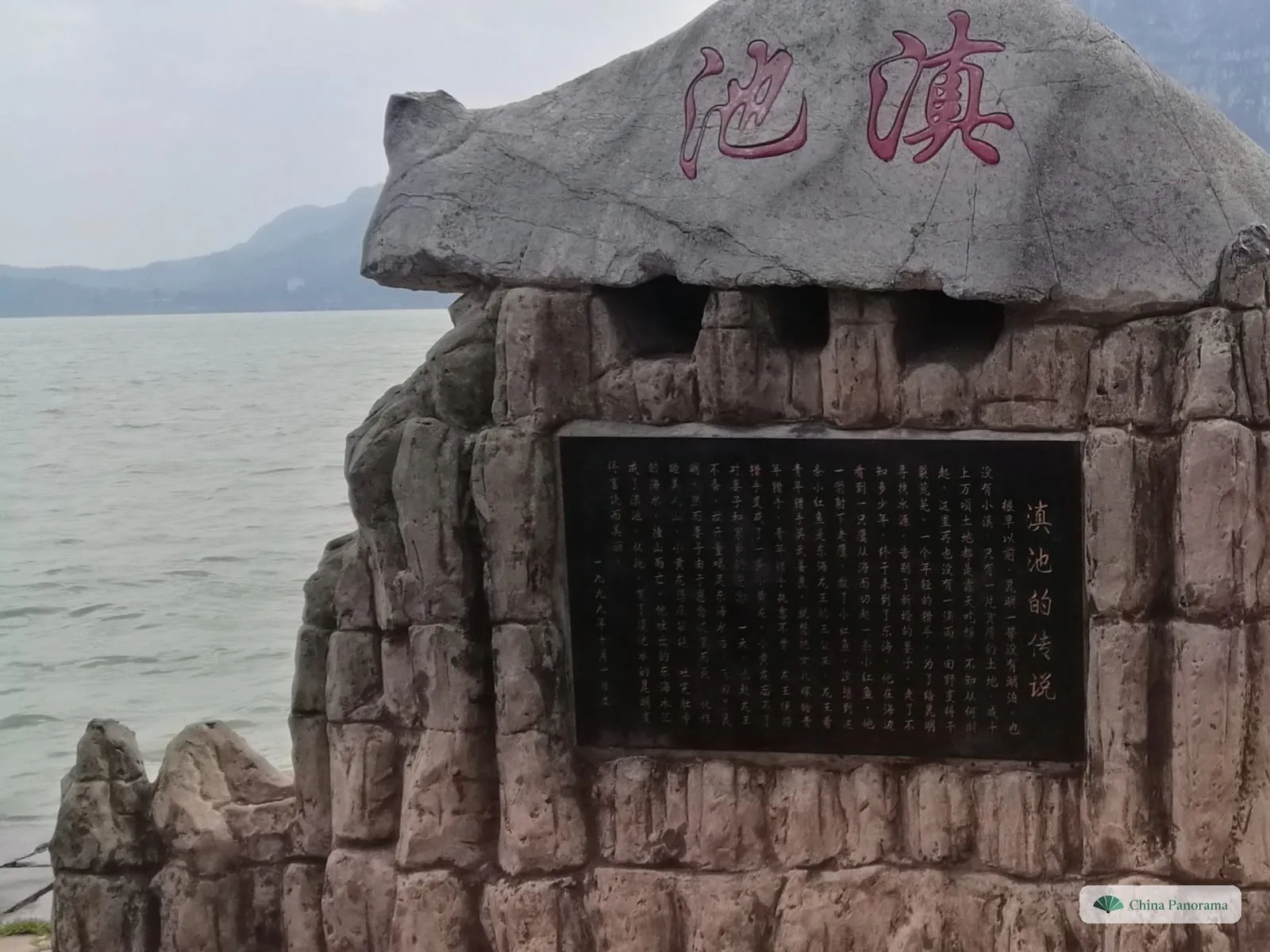 A grey boulder carved with two red characters on top of an artificial rock base holding a black plaque of engraved text, a wide lake and hills behind