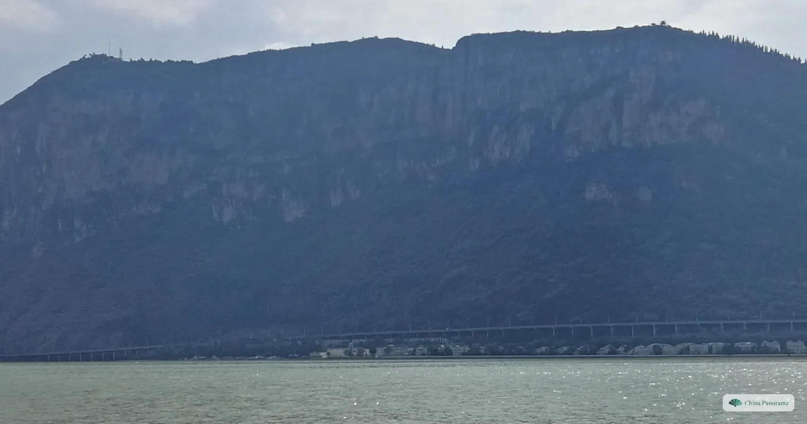 A wide lake under a long mountain ridge whose face drops in grey cliffs, a line of buildings along the far shore