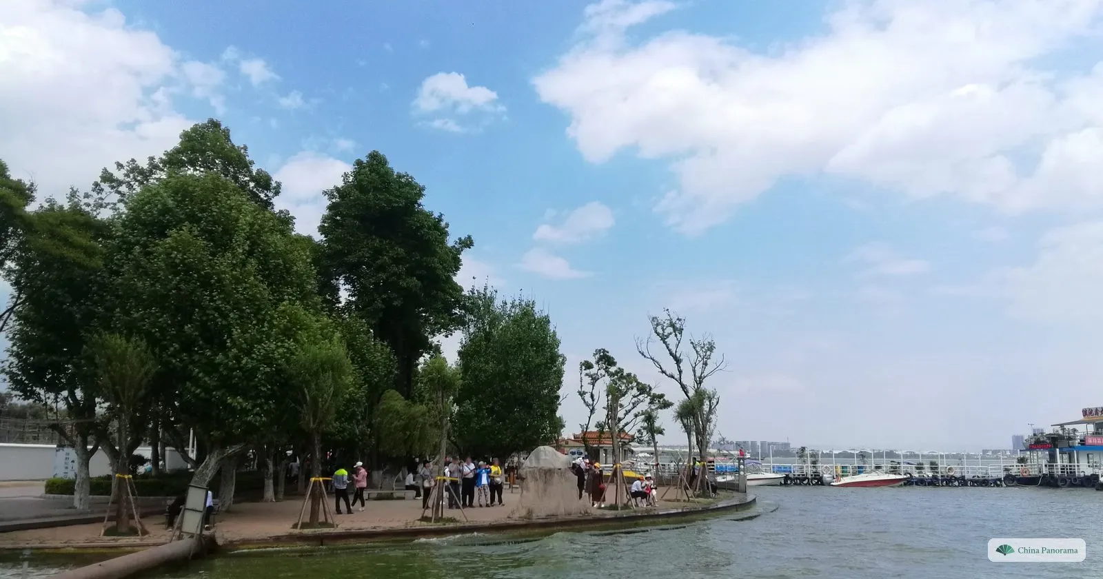A curving waterfront with trees, a carved boulder and people, speedboats and a pier on a wide stretch of water
