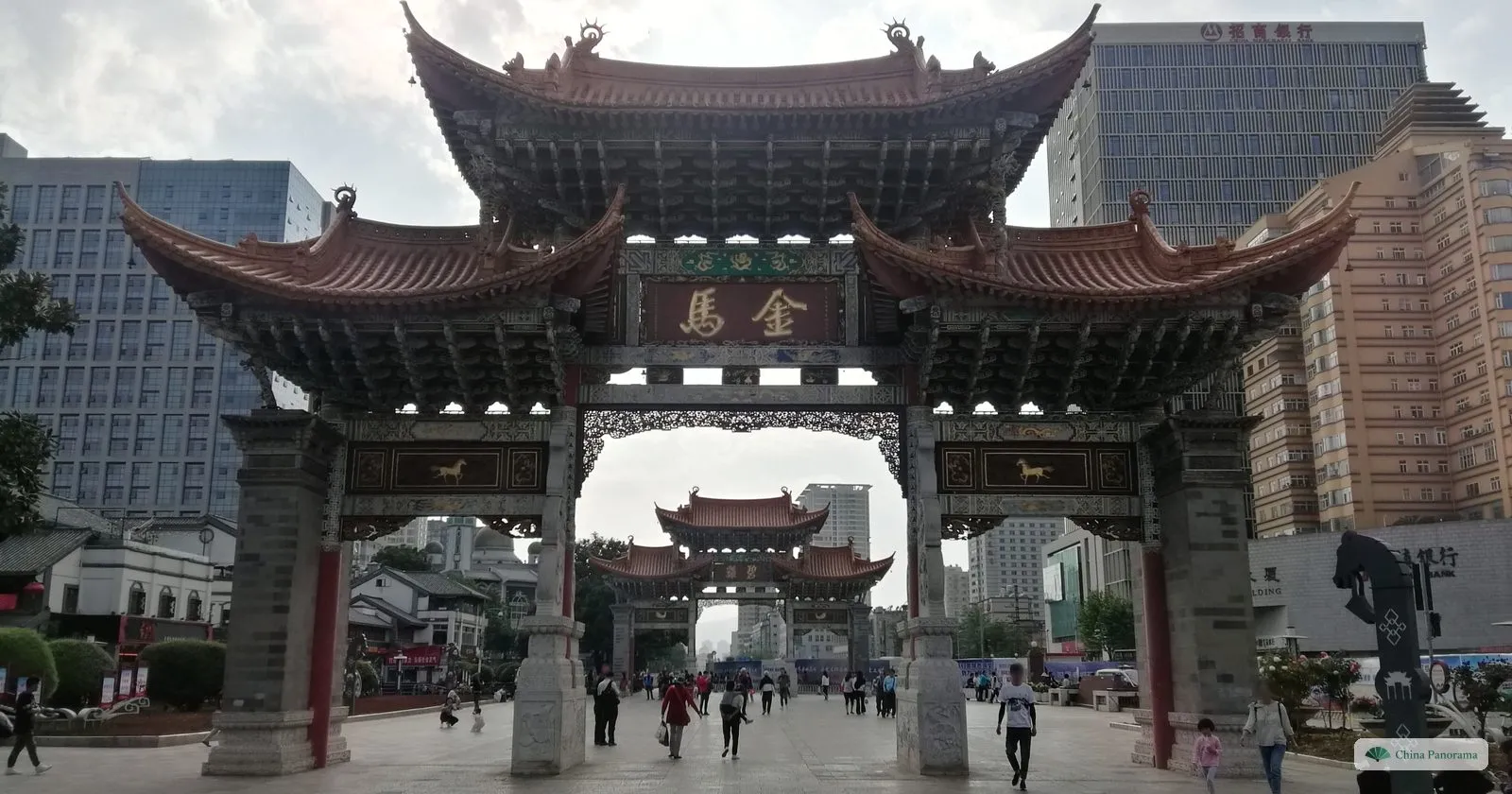 A huge wooden archway with tiered roofs and a plaque of two gold characters, and through its central opening a second, similar archway further along a pedestrian street