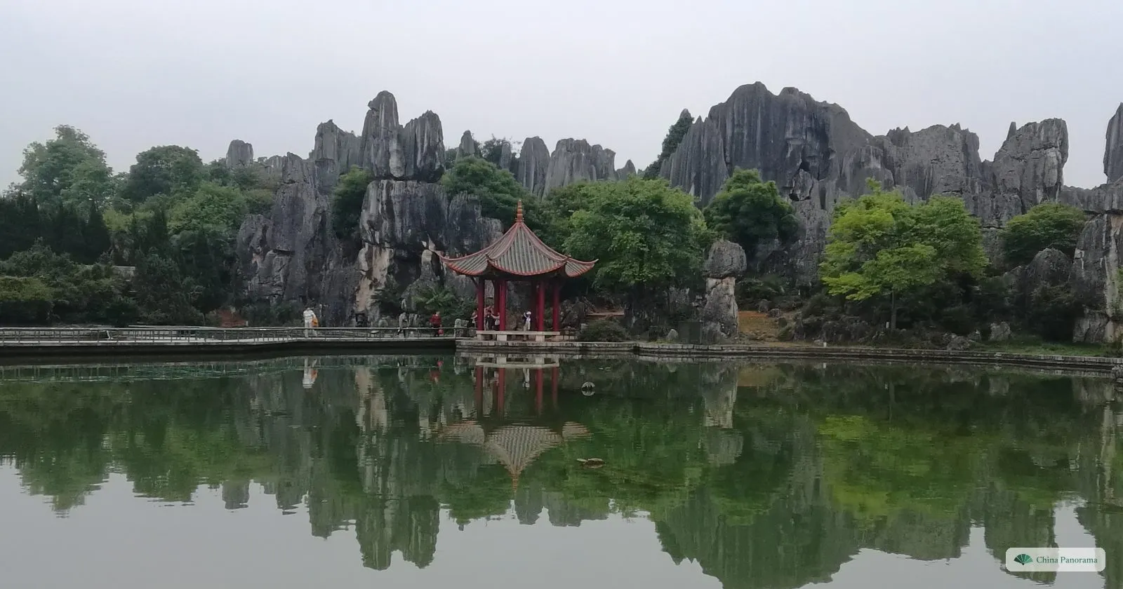 A red pavilion and a jagged line of grey rocks reflected in a calm pond under an overcast sky
