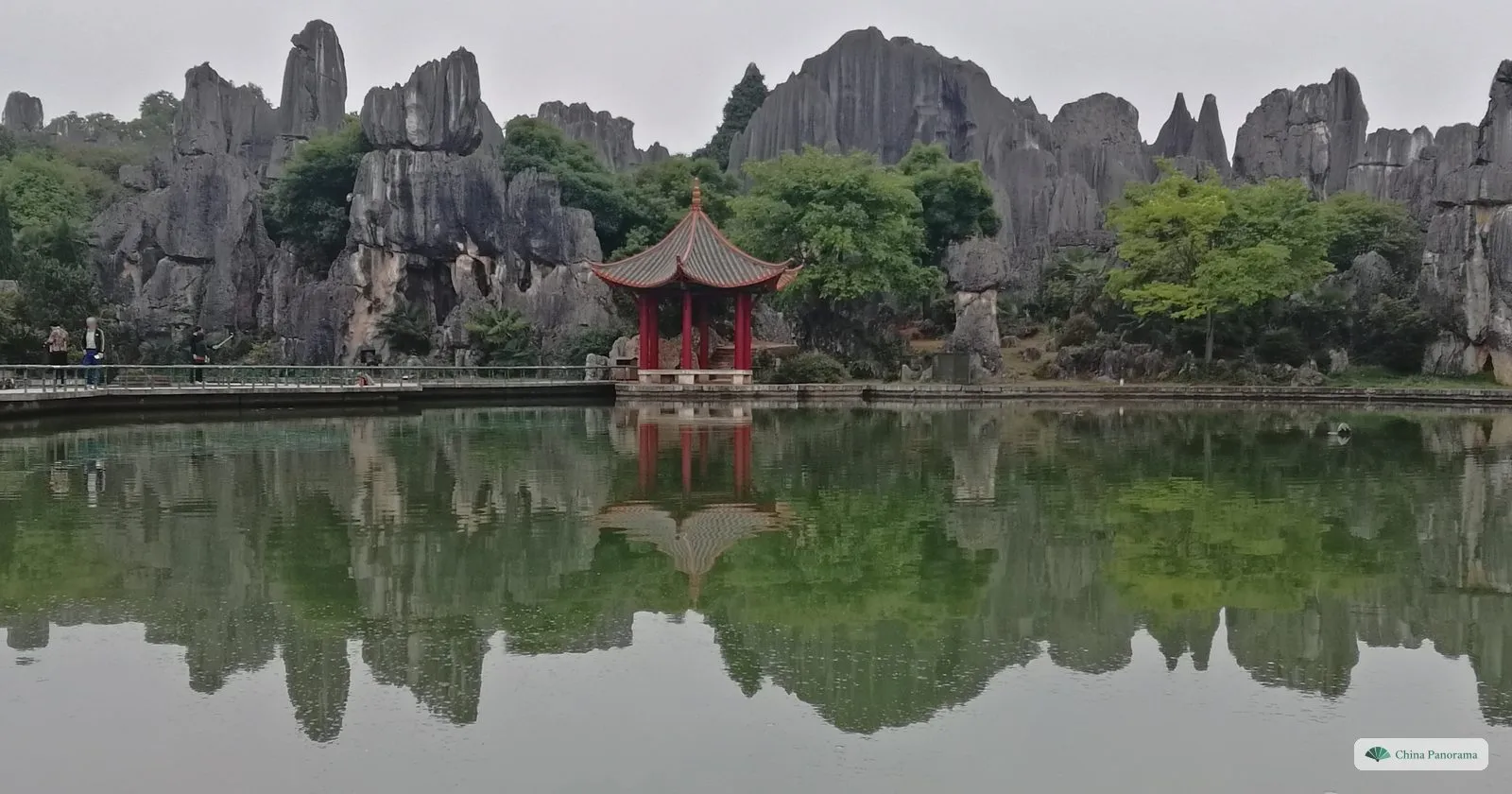 A red pavilion on the edge of a still pond, grey rock pinnacles and trees behind it and the whole scene mirrored in the water