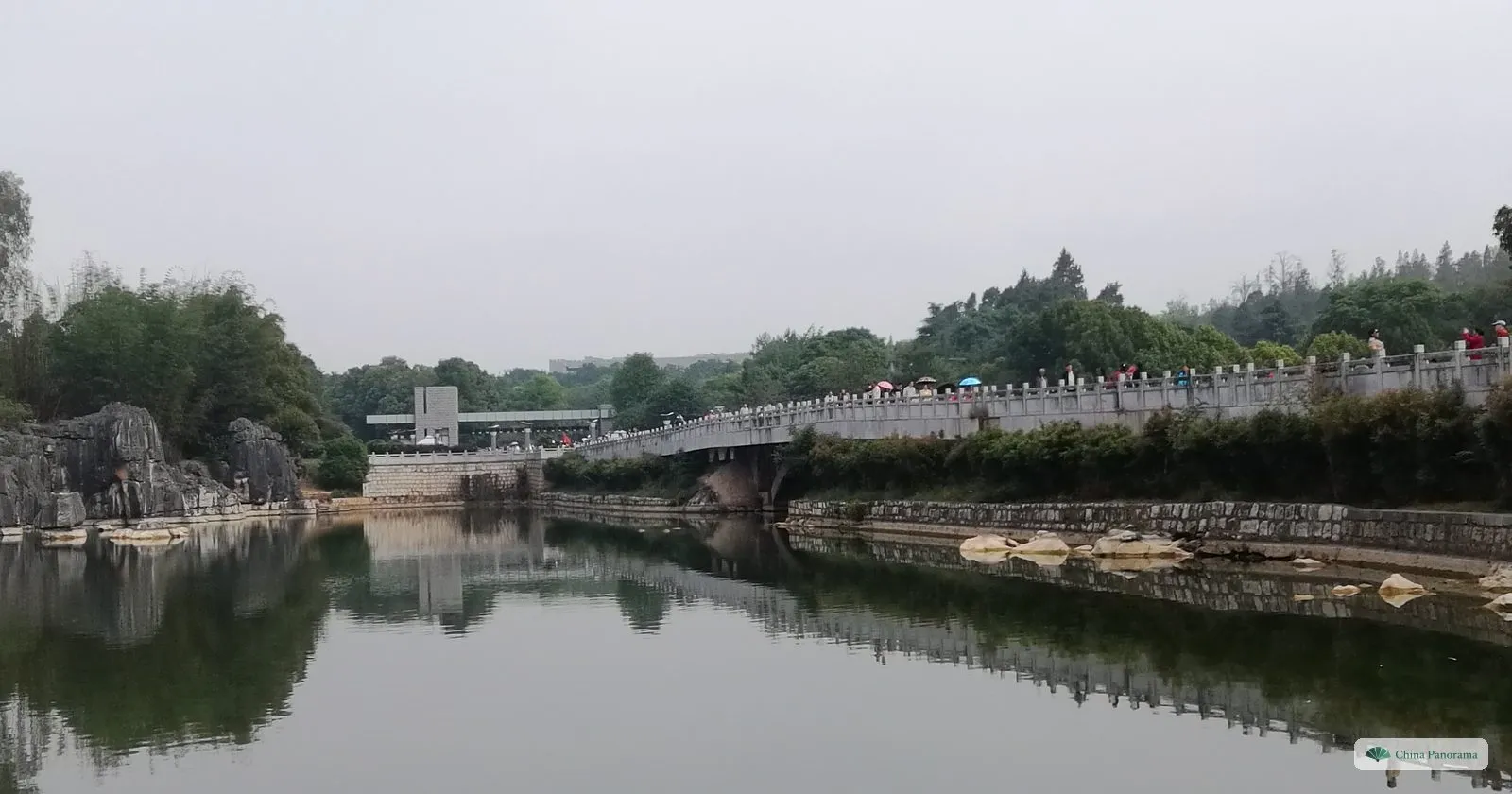 A long balustraded bridge across a calm lake, rocks at the water’s edge on the left, a low building at the far end