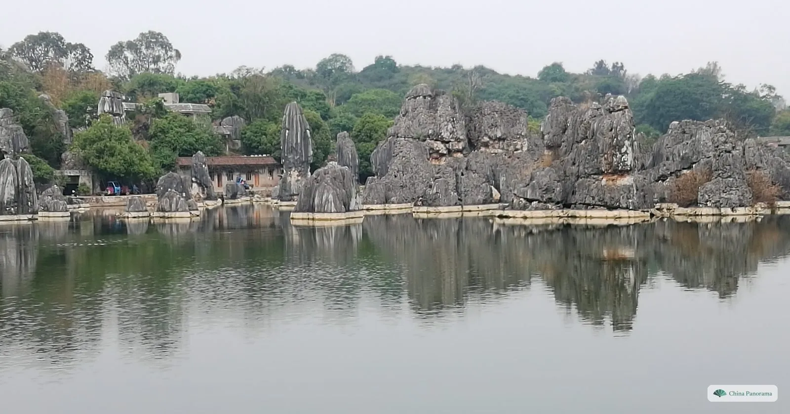 Grey limestone formations along the shore of a calm lake under a white sky, their reflections in the water