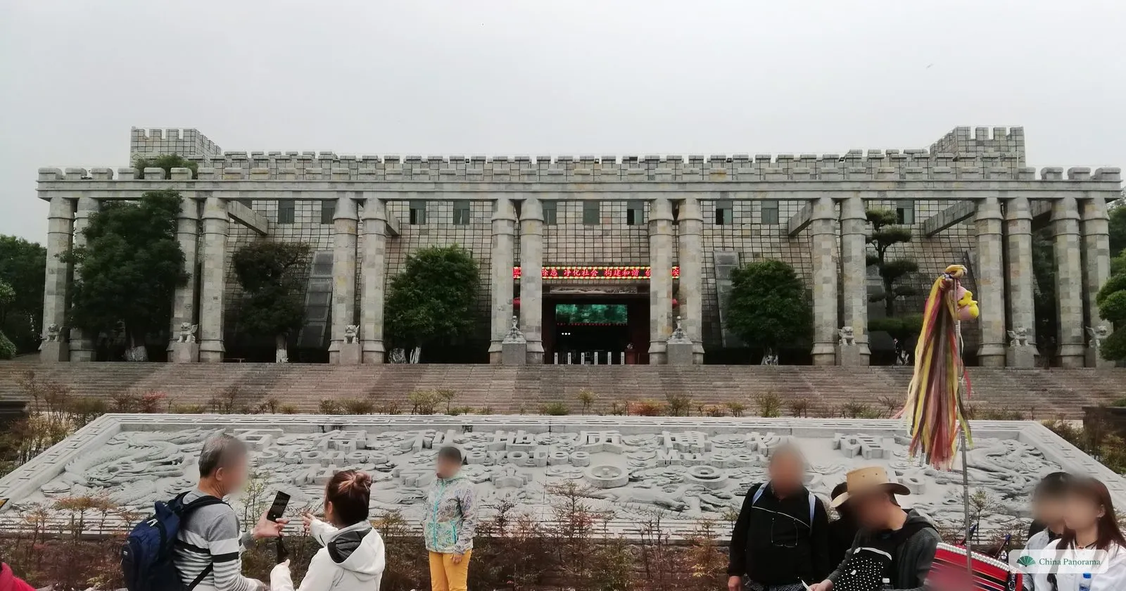 A large grey building with square columns and battlements at the top of wide steps, a carved stone relief in front and tourists taking photos